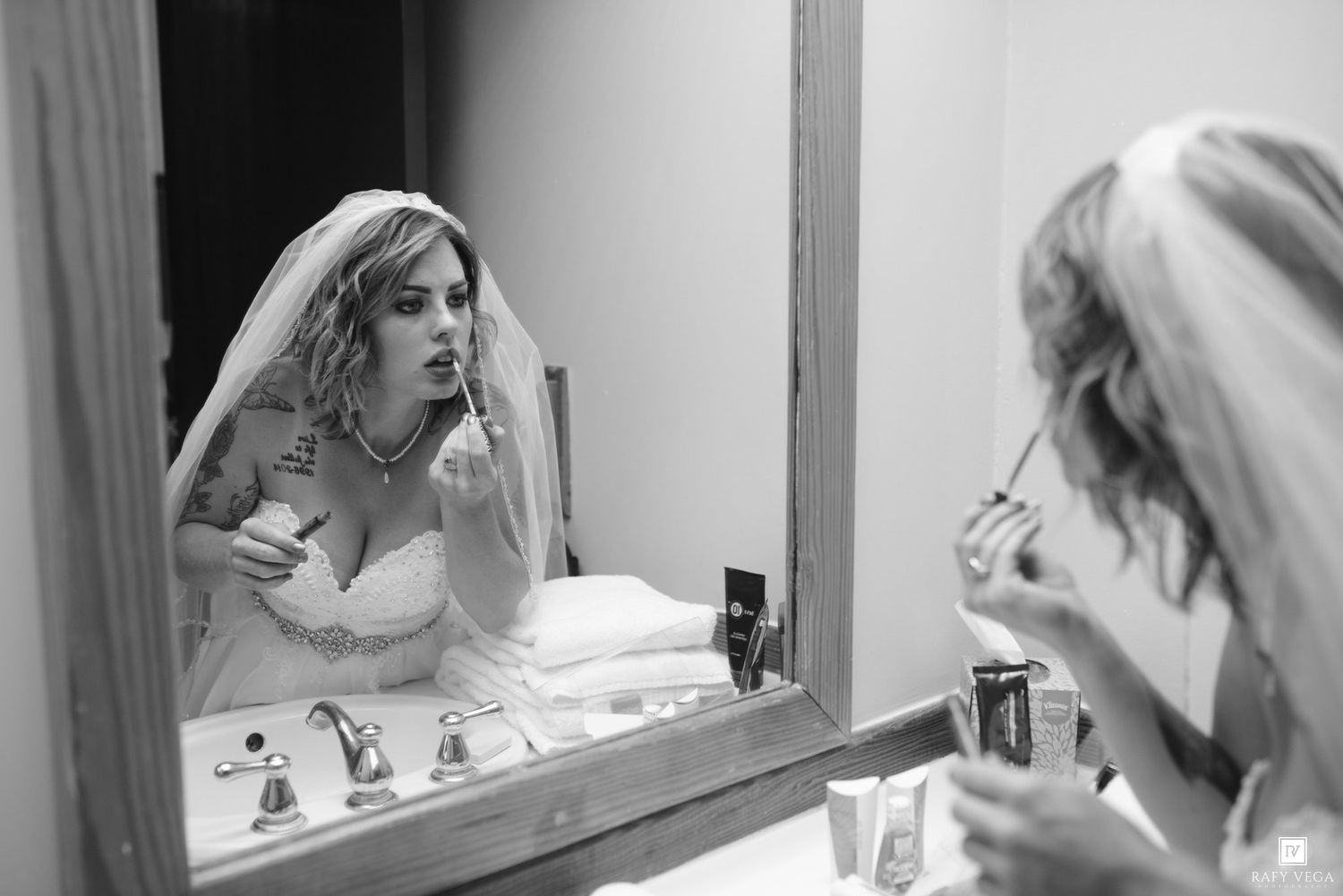 Black and white photo of a bride in veil applying lipstick at a bathroom mirror on her wedding day.