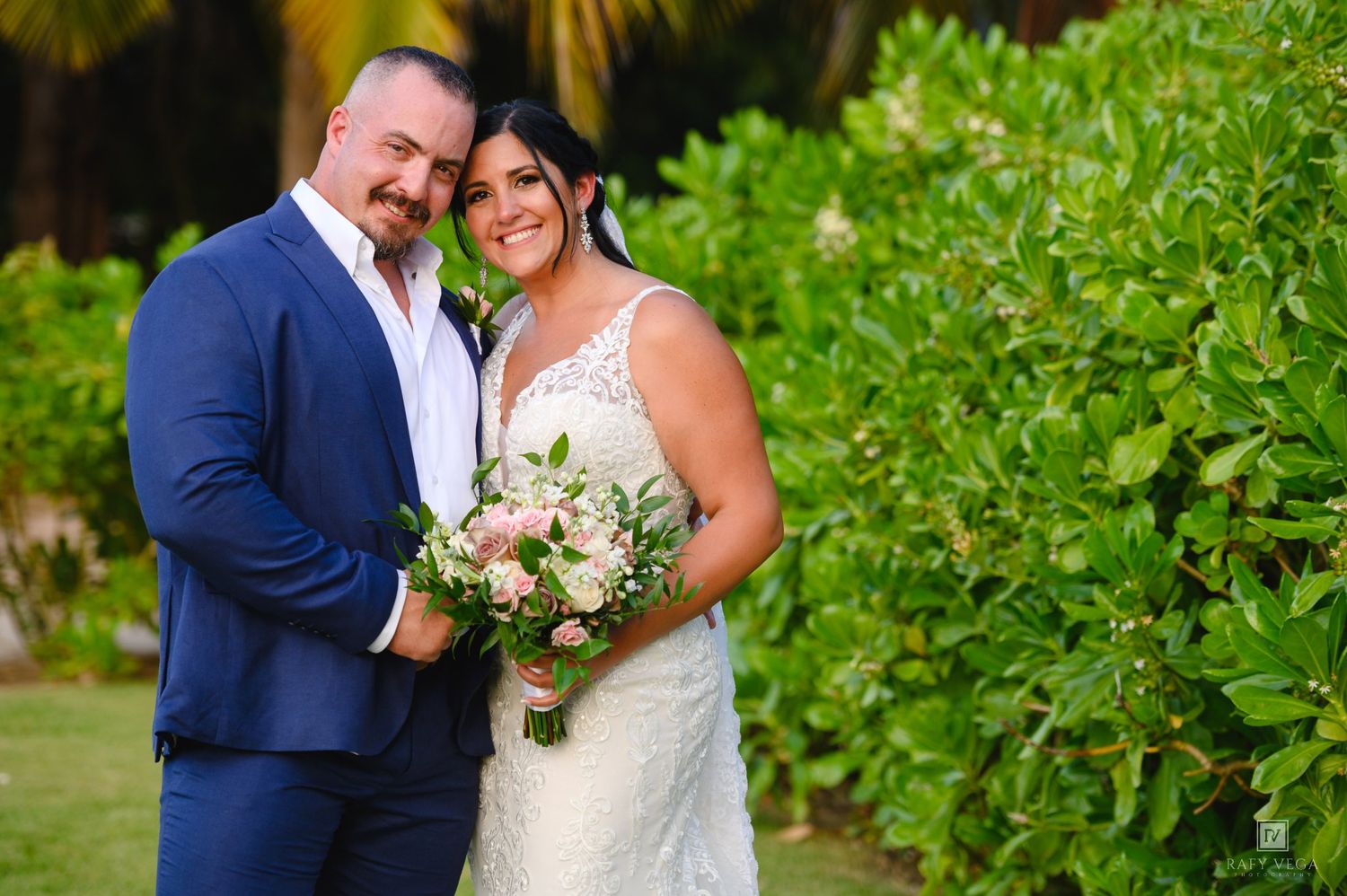 Bride and groom pose together outdoors, she holds pink bouquet, he wears blue suit, lush greenery background.