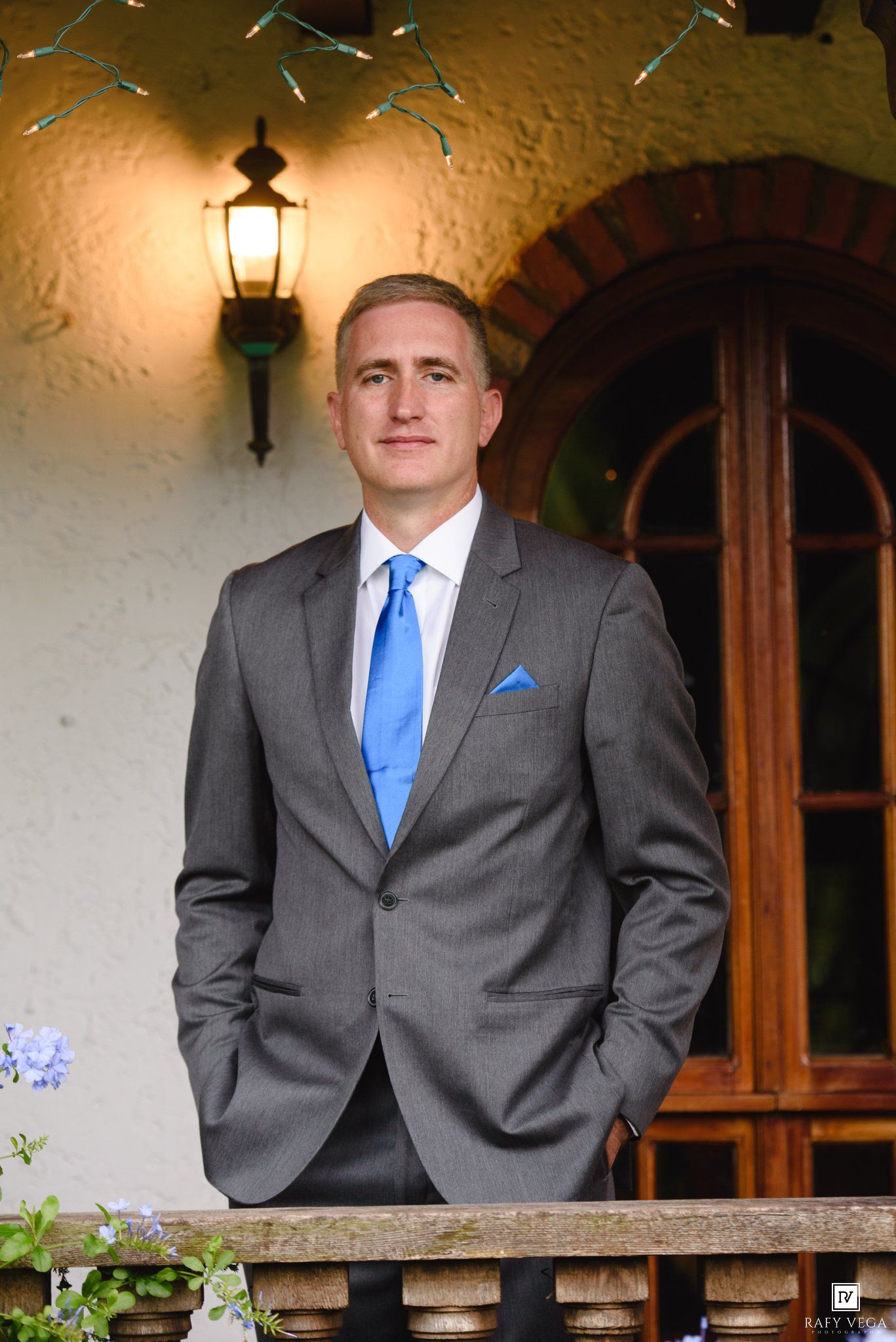 Man in gray suit with blue tie and pocket square standing on a balcony near an arched wooden door.