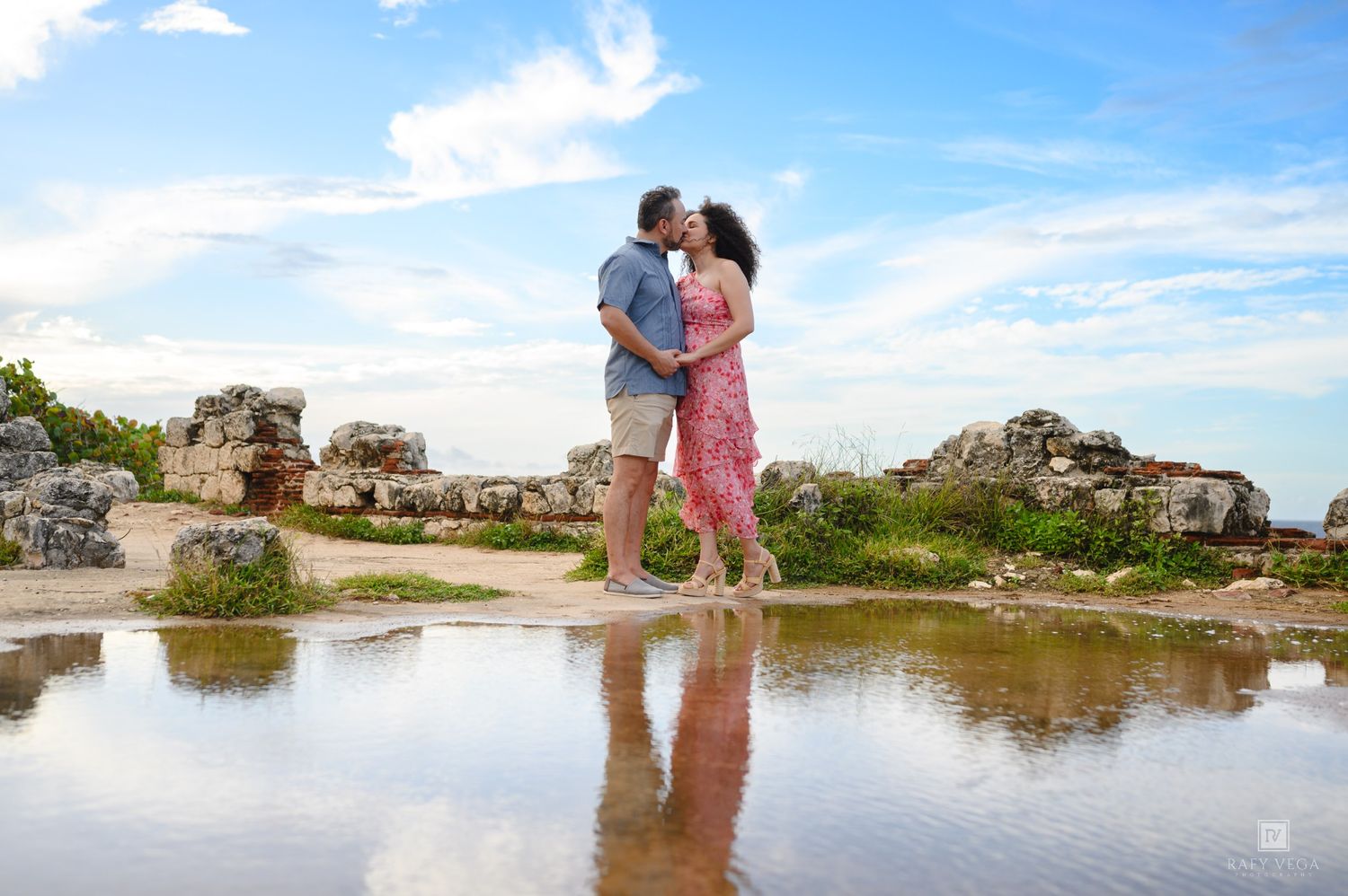 Couple kissing near ancient ruins with reflective water puddle under a blue sky with clouds.