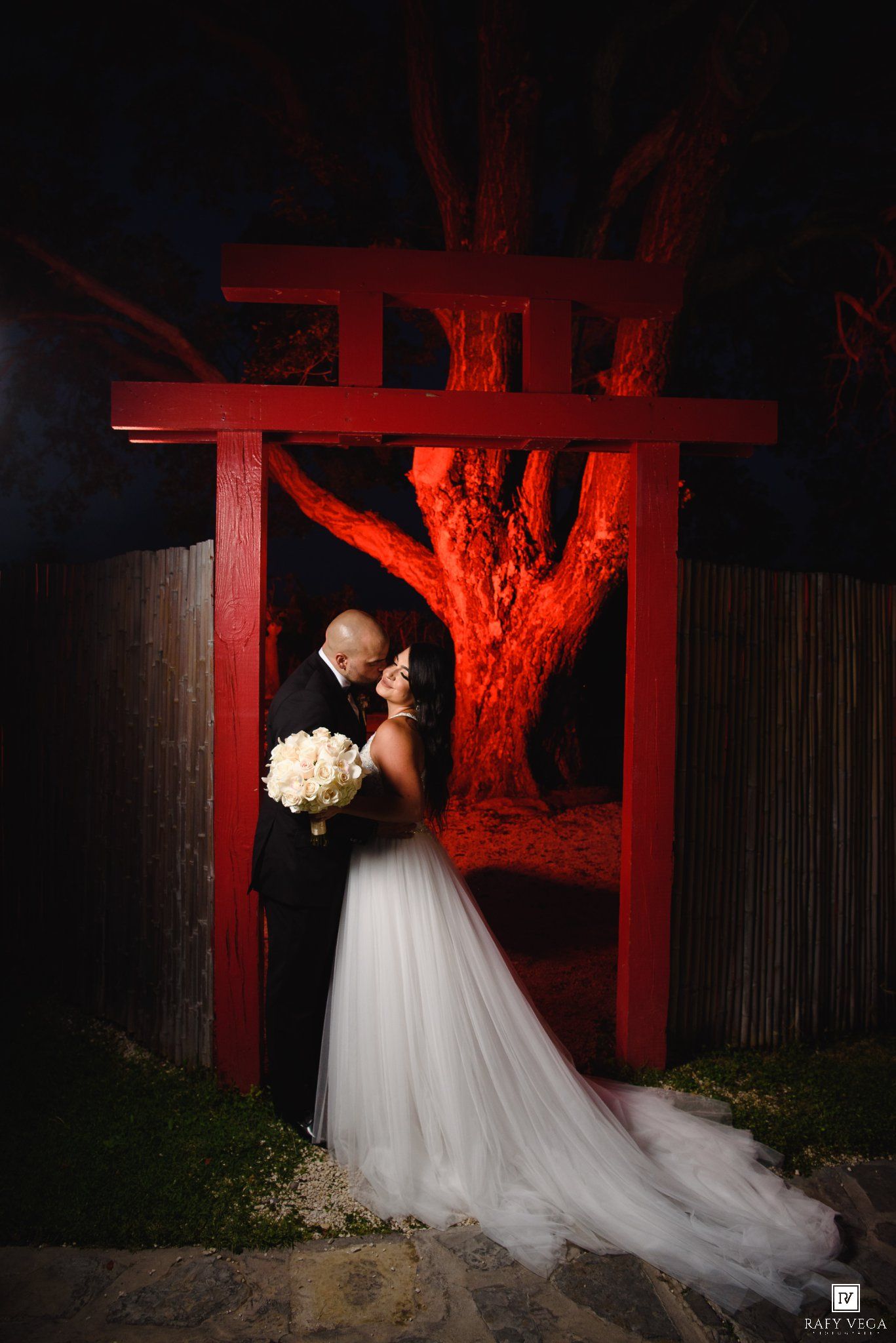 Bride and groom share a romantic kiss under a red torii gate at night, with a glowing tree backdrop.