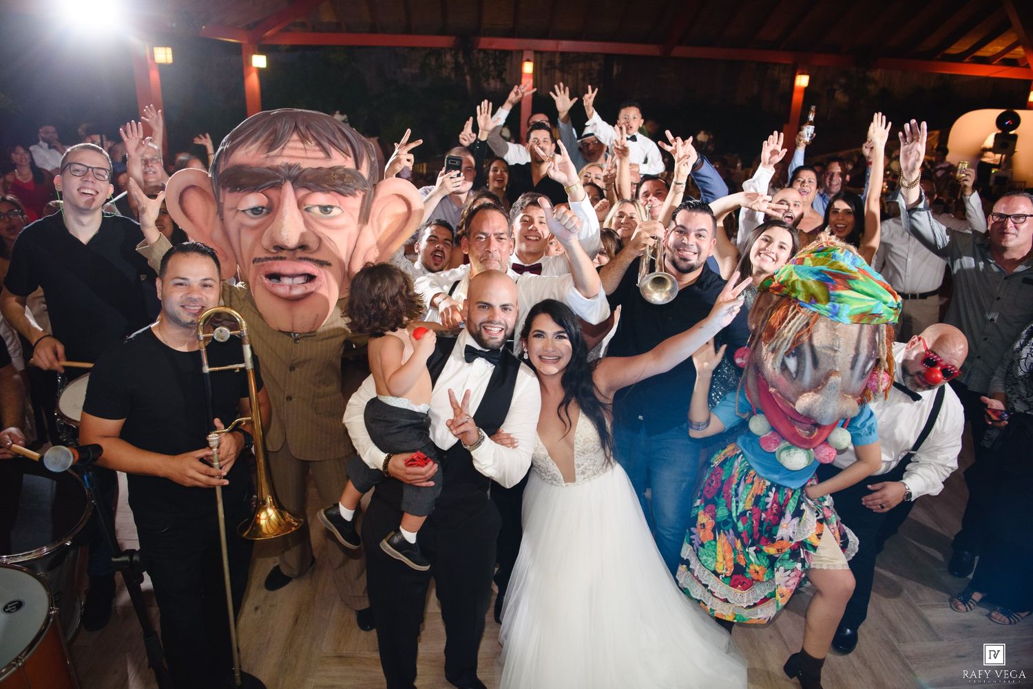 Wedding couple poses with guests and giant puppet mascots at a lively outdoor wedding celebration party.
