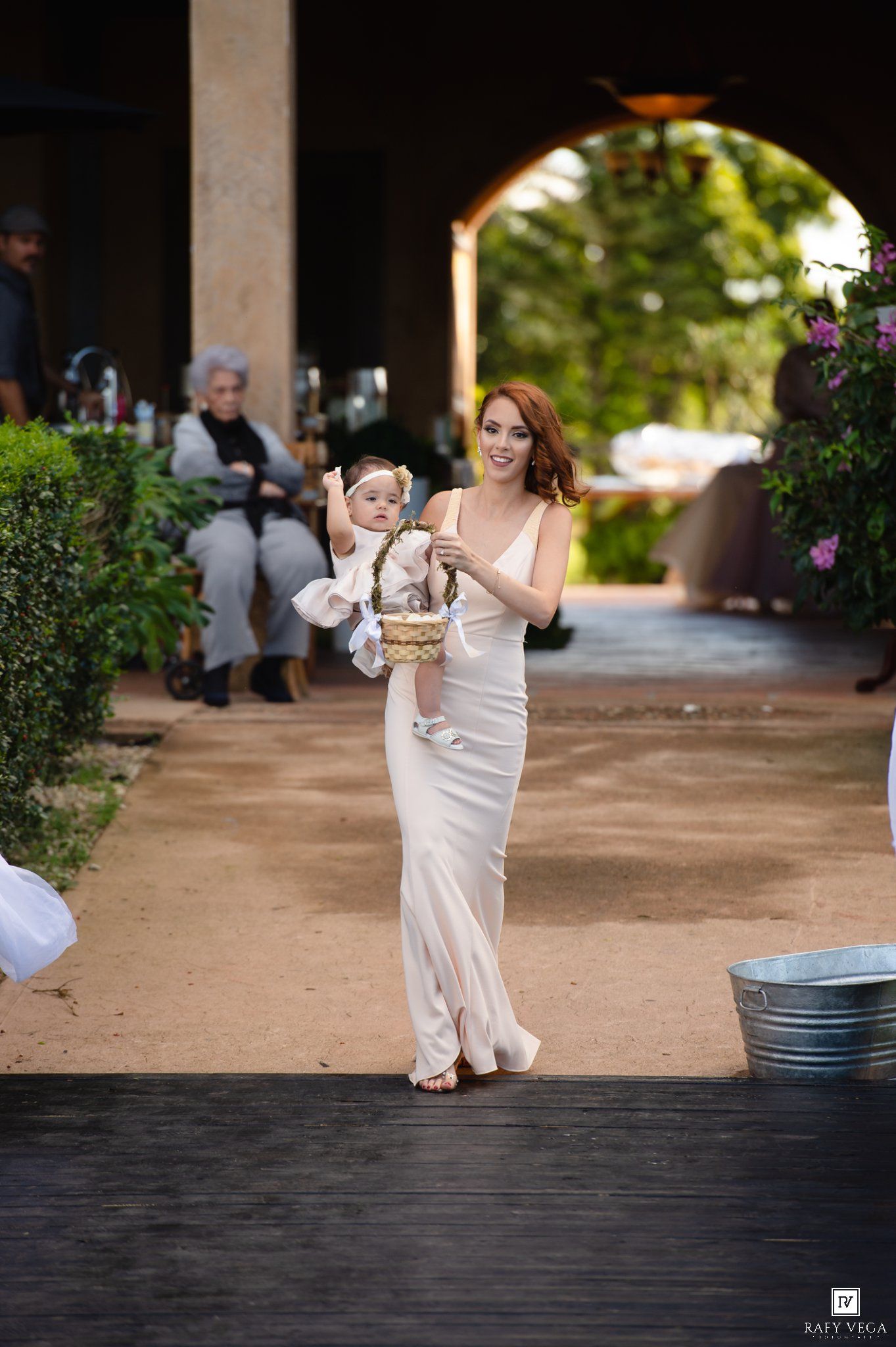 A woman in a white gown carries a baby with a flower basket down an outdoor wedding aisle.