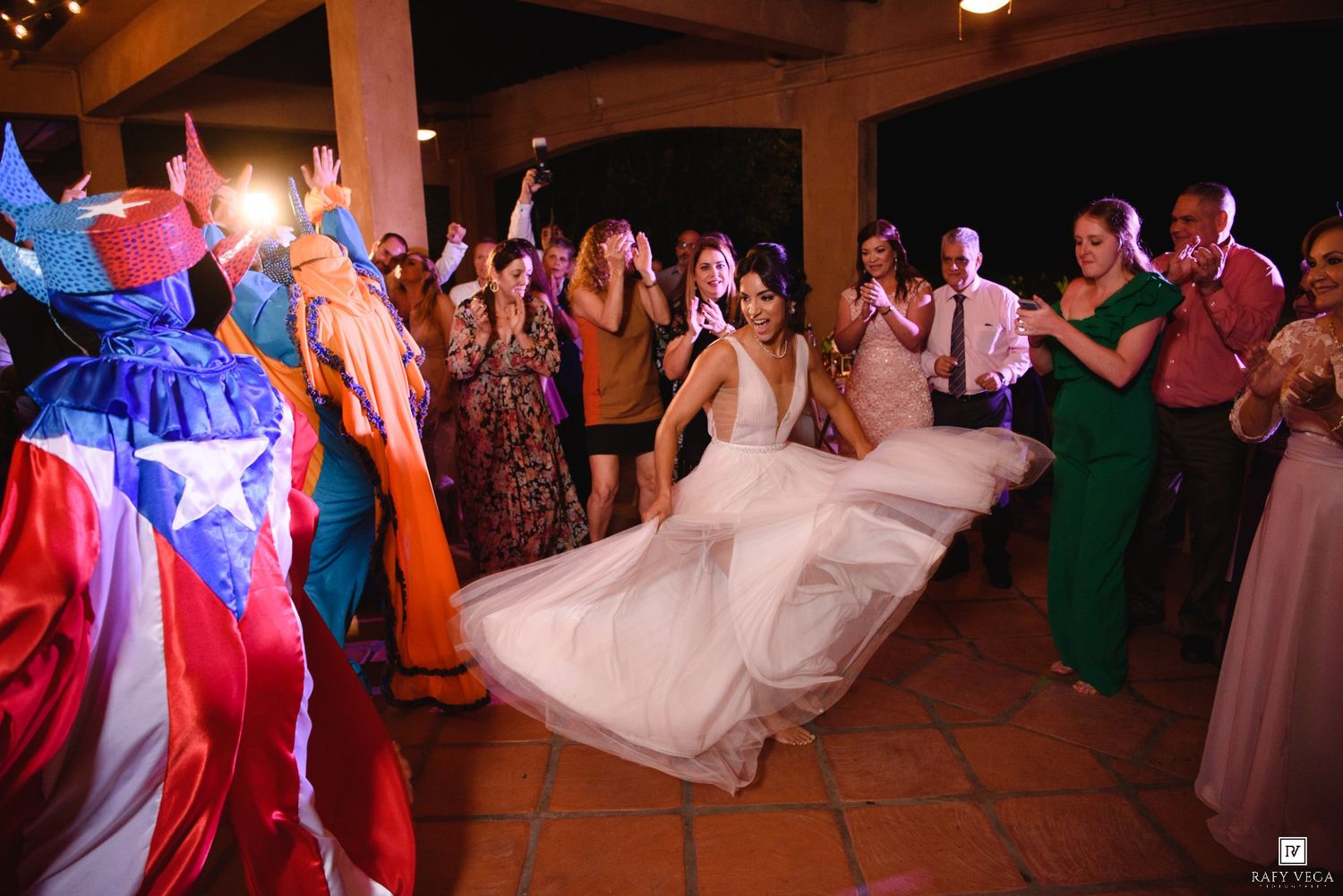 Bride dances joyfully surrounded by cheering guests in colorful outfits at a festive wedding reception.