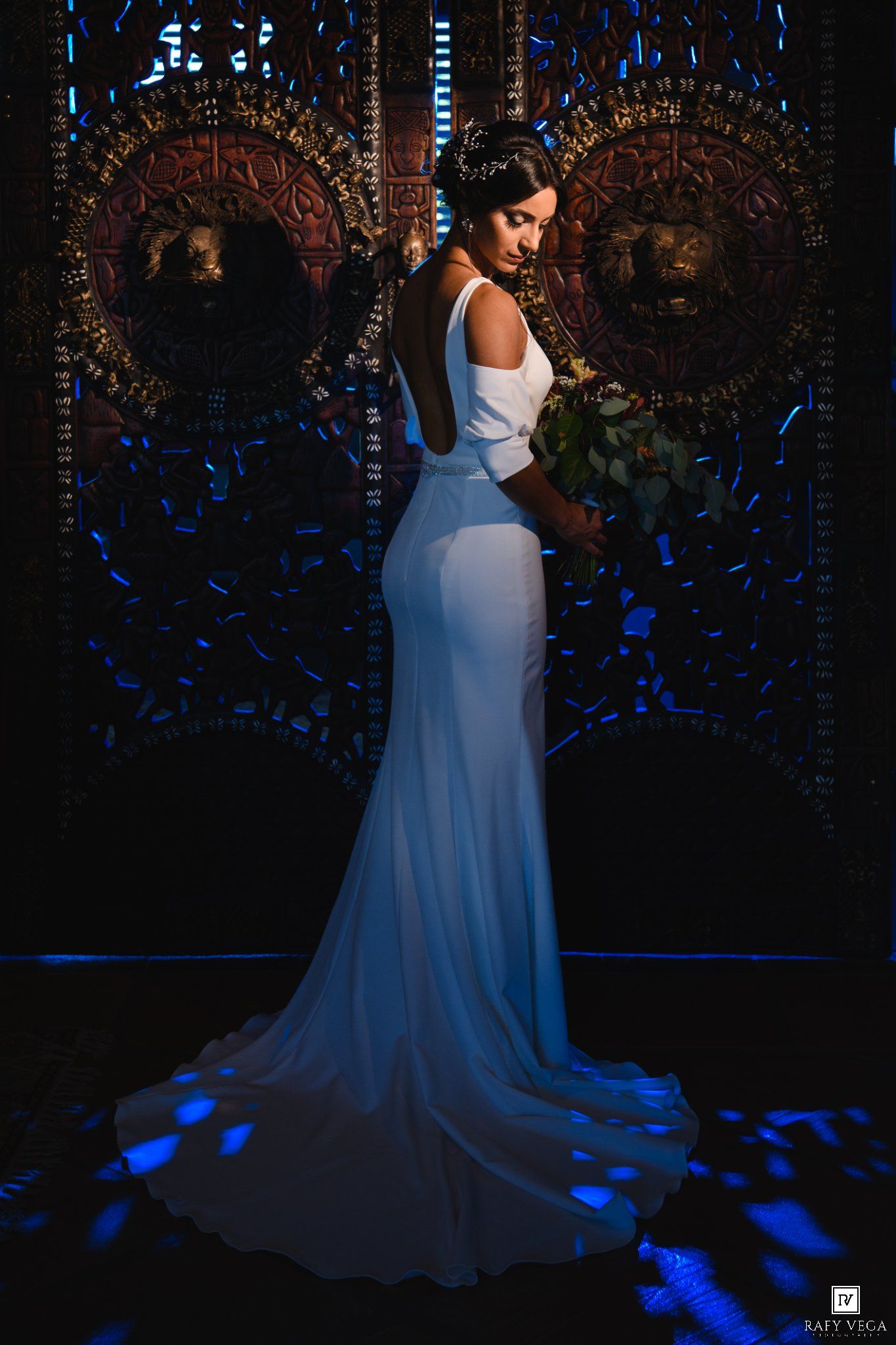 Bride in elegant white backless gown poses dramatically against ornate dark backdrop with blue lighting.