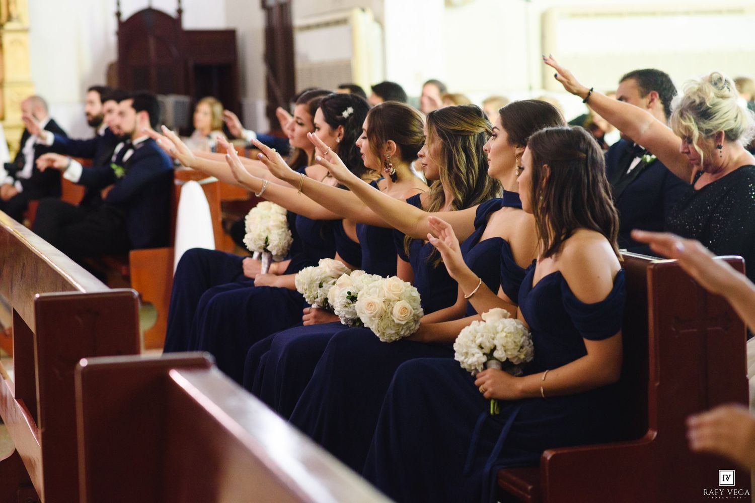 Bridesmaids in navy blue dresses holding white bouquets seated in church pews during a wedding ceremony.
