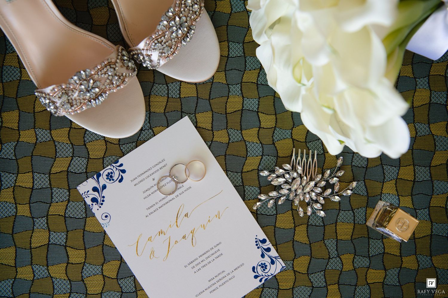 Wedding flat lay with embellished bridal heels, invitation, rings, pearl hair comb, and white roses on patterned fabric.