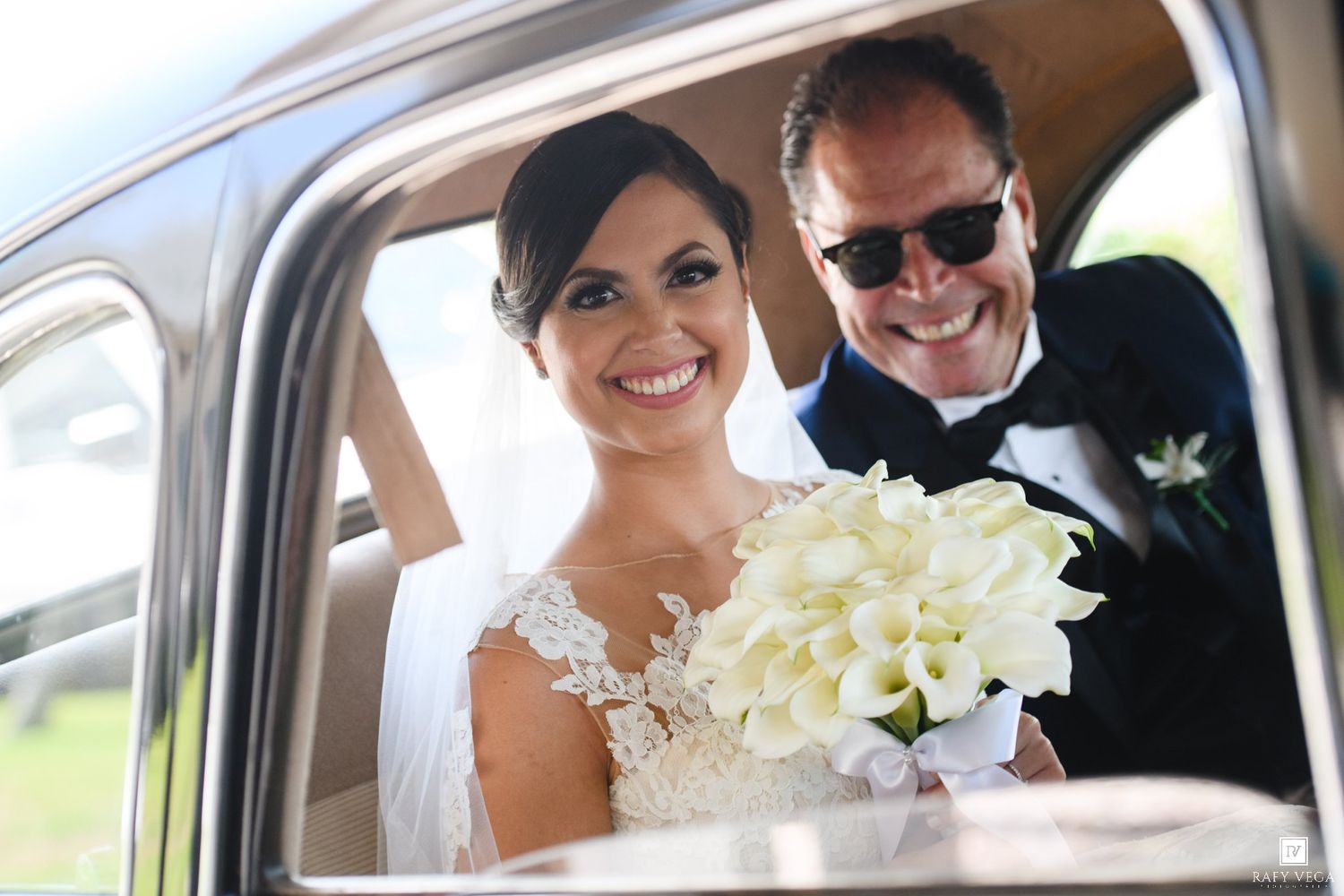 Smiling bride holding white calla lily bouquet sits in vintage car with suited man wearing sunglasses on wedding day.