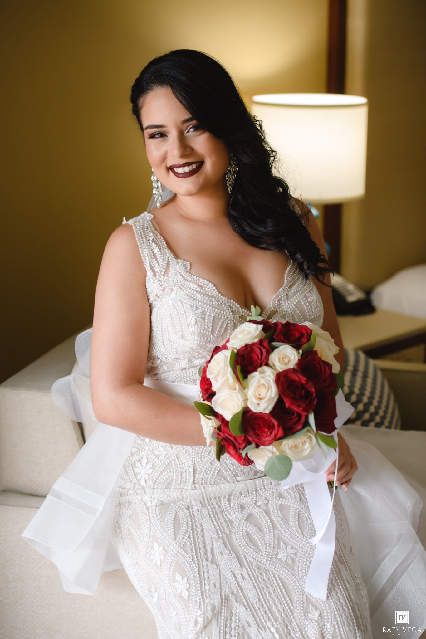 Smiling bride in beaded white gown holding red and white rose bouquet, seated indoors with elegant earrings.