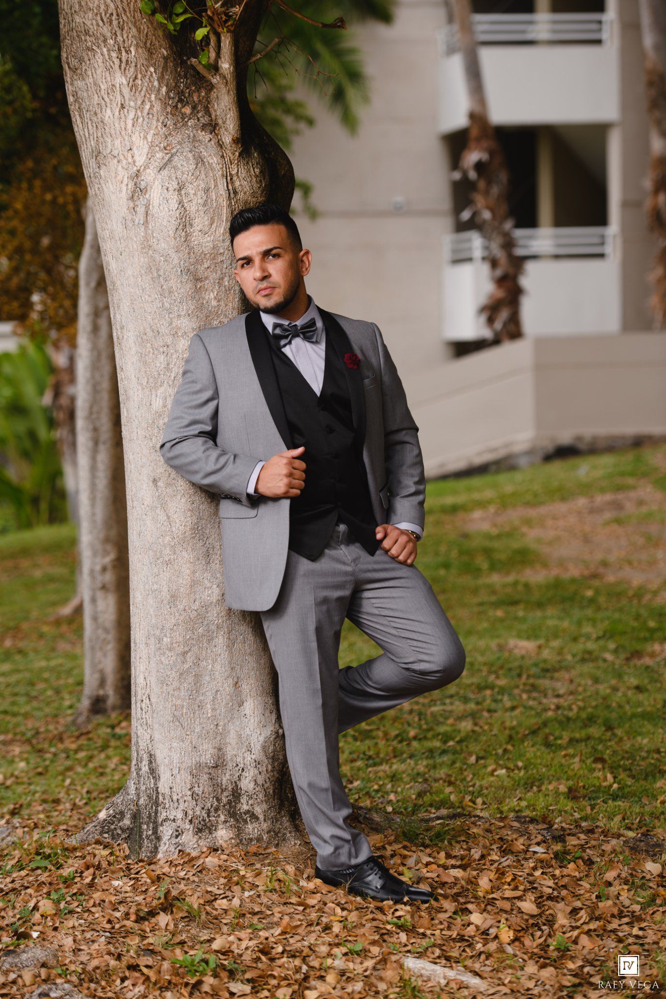 Dapper groom in gray suit with black vest and bow tie leaning against tree outdoors.