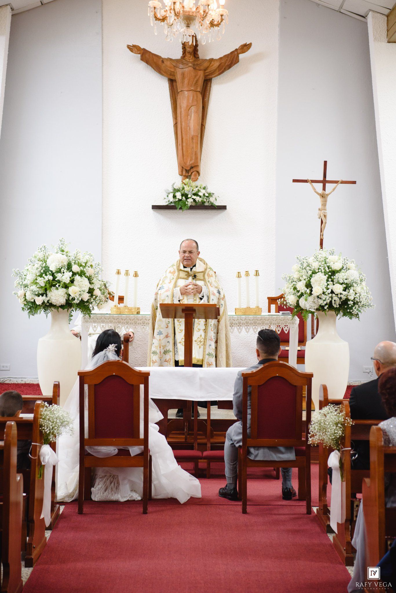 Couple exchanging vows at a Catholic church wedding ceremony with white floral arrangements and a priest at the altar.