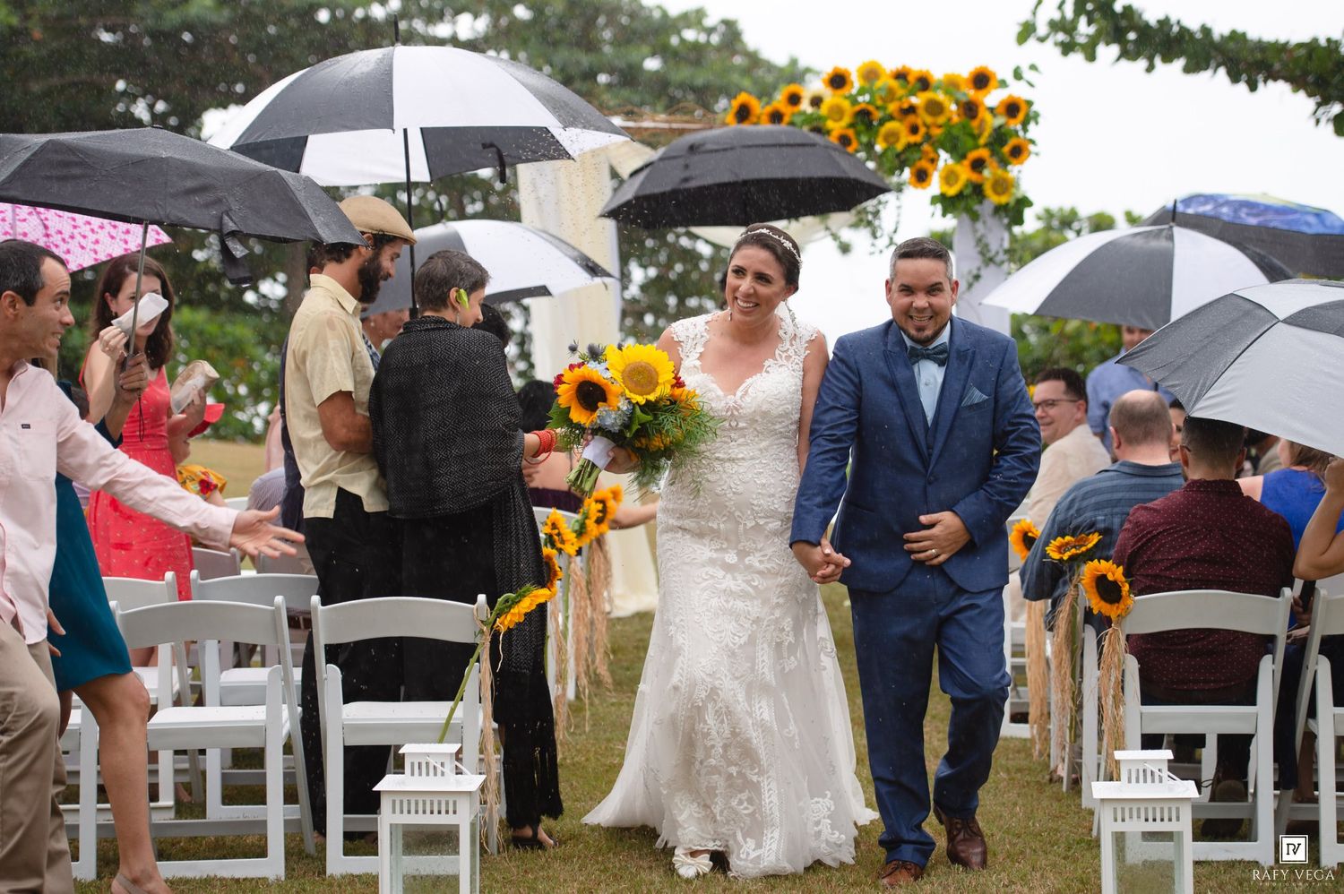 Bride and groom walk down aisle in outdoor wedding ceremony with sunflower arch and guests holding black umbrellas.