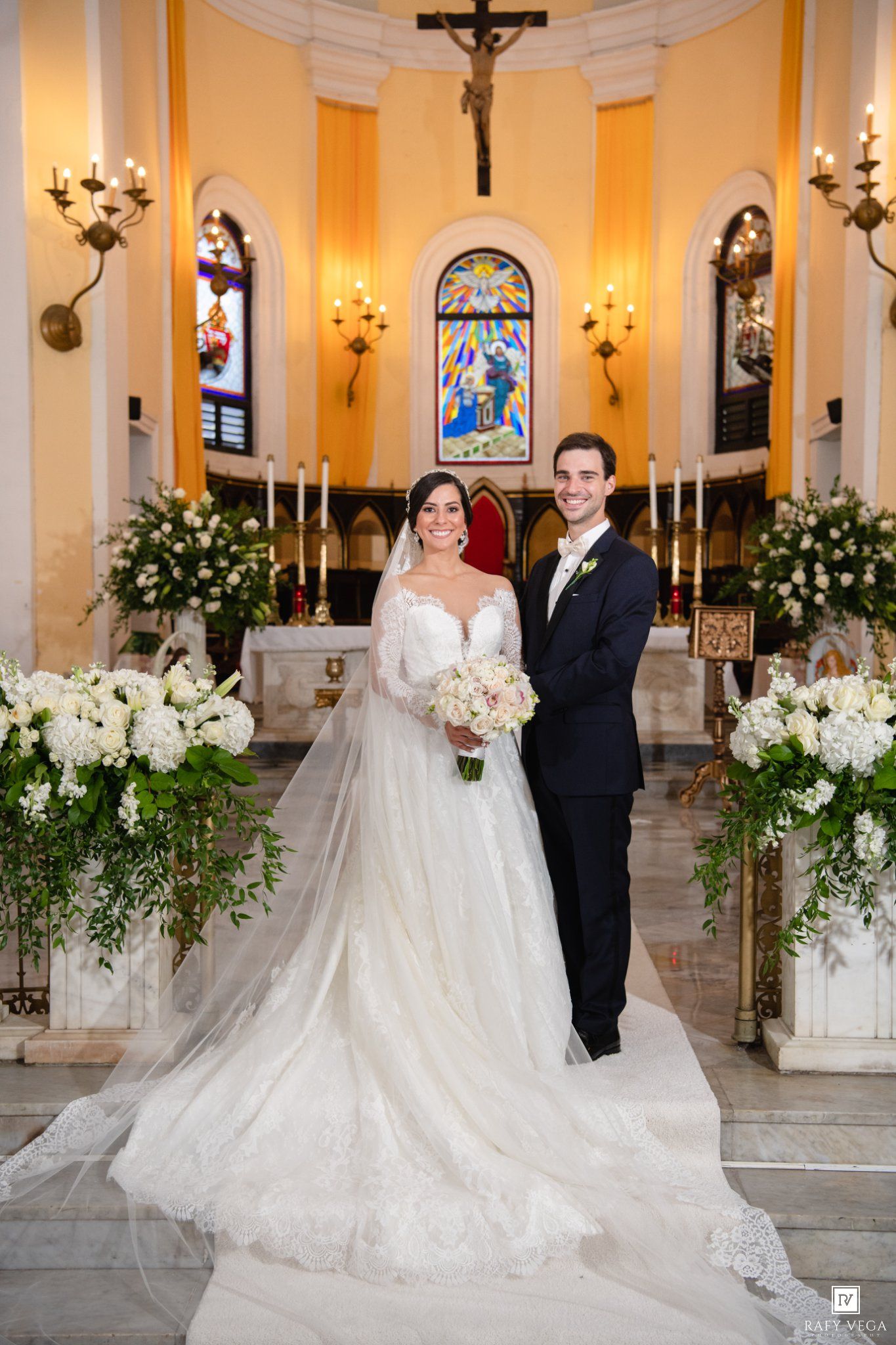 Bride and groom pose together at church altar surrounded by white flowers after their wedding ceremony.