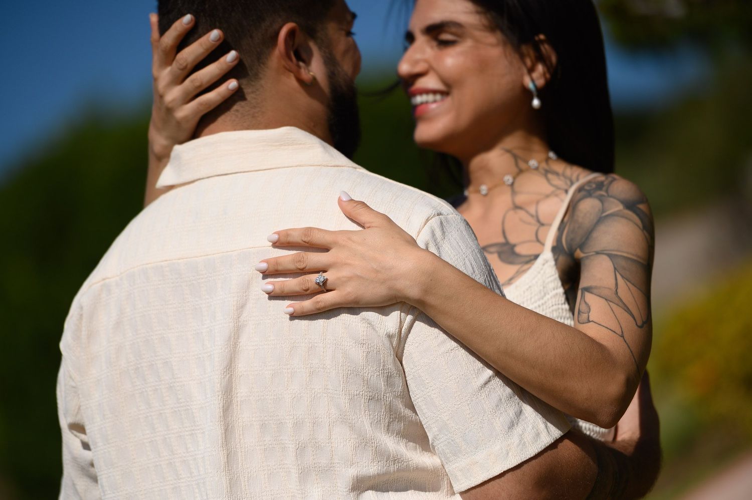 Smiling tattooed woman embraces a man outdoors, showcasing an engagement ring on her finger.