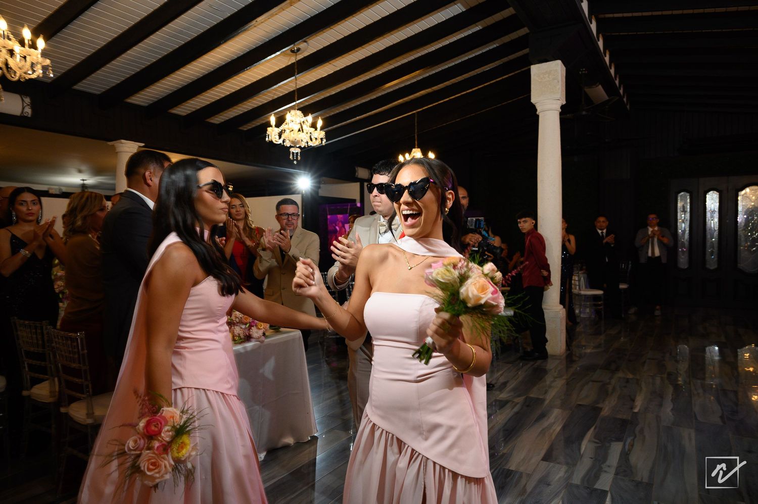 Two bridesmaids in blush pink dresses and sunglasses hold bouquets and laugh while dancing at a wedding reception as guests watch.