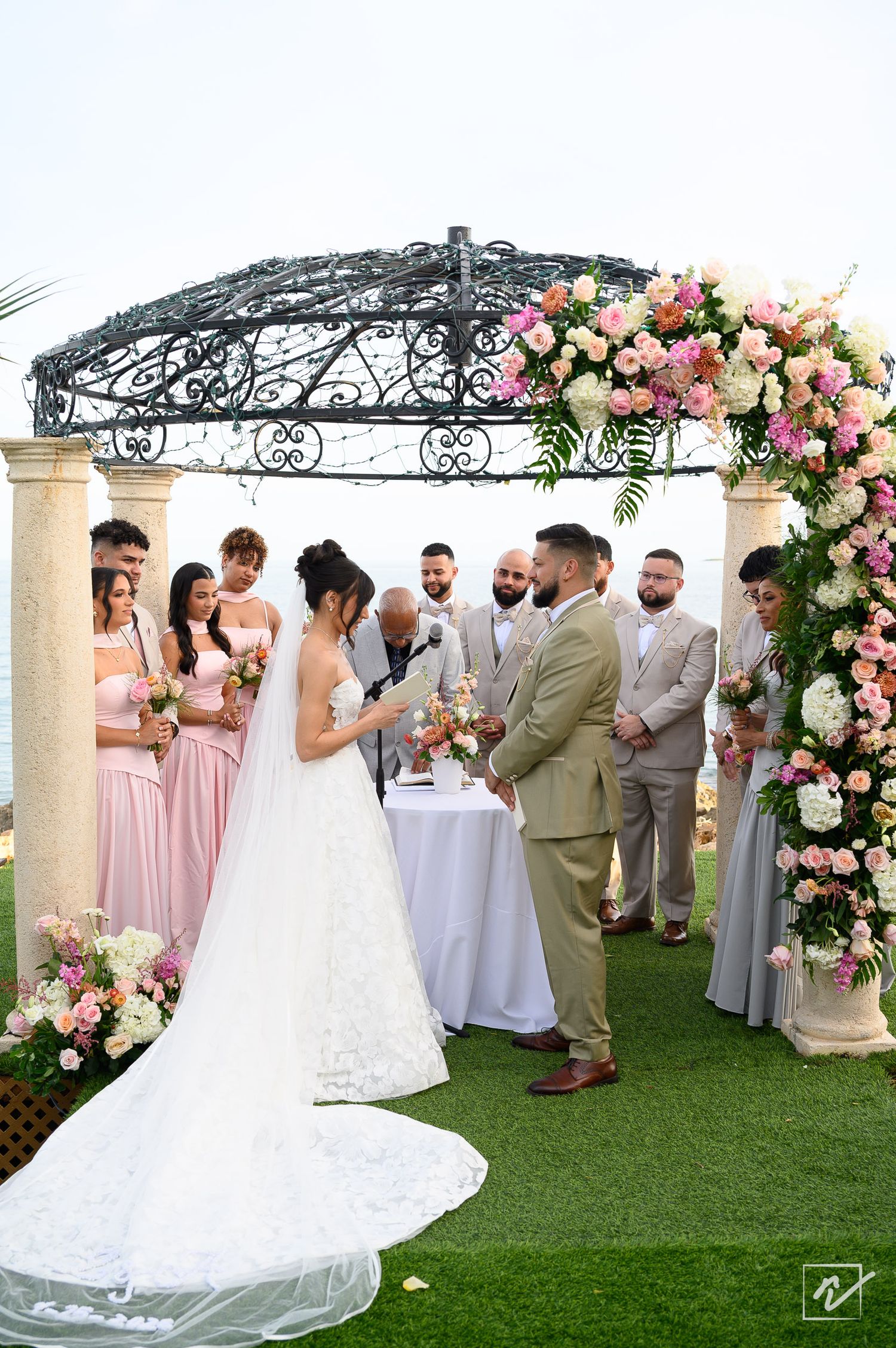 Bride in a long lace gown and groom in an olive suit exchange vows under an ornate gazebo with pink floral arrangements and wedding party.