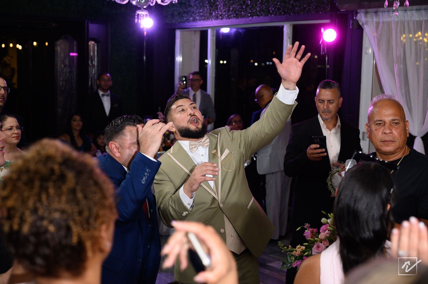 Groom in a green suit raises his hand while guests film with phones during a lively wedding reception in a softly lit ballroom.