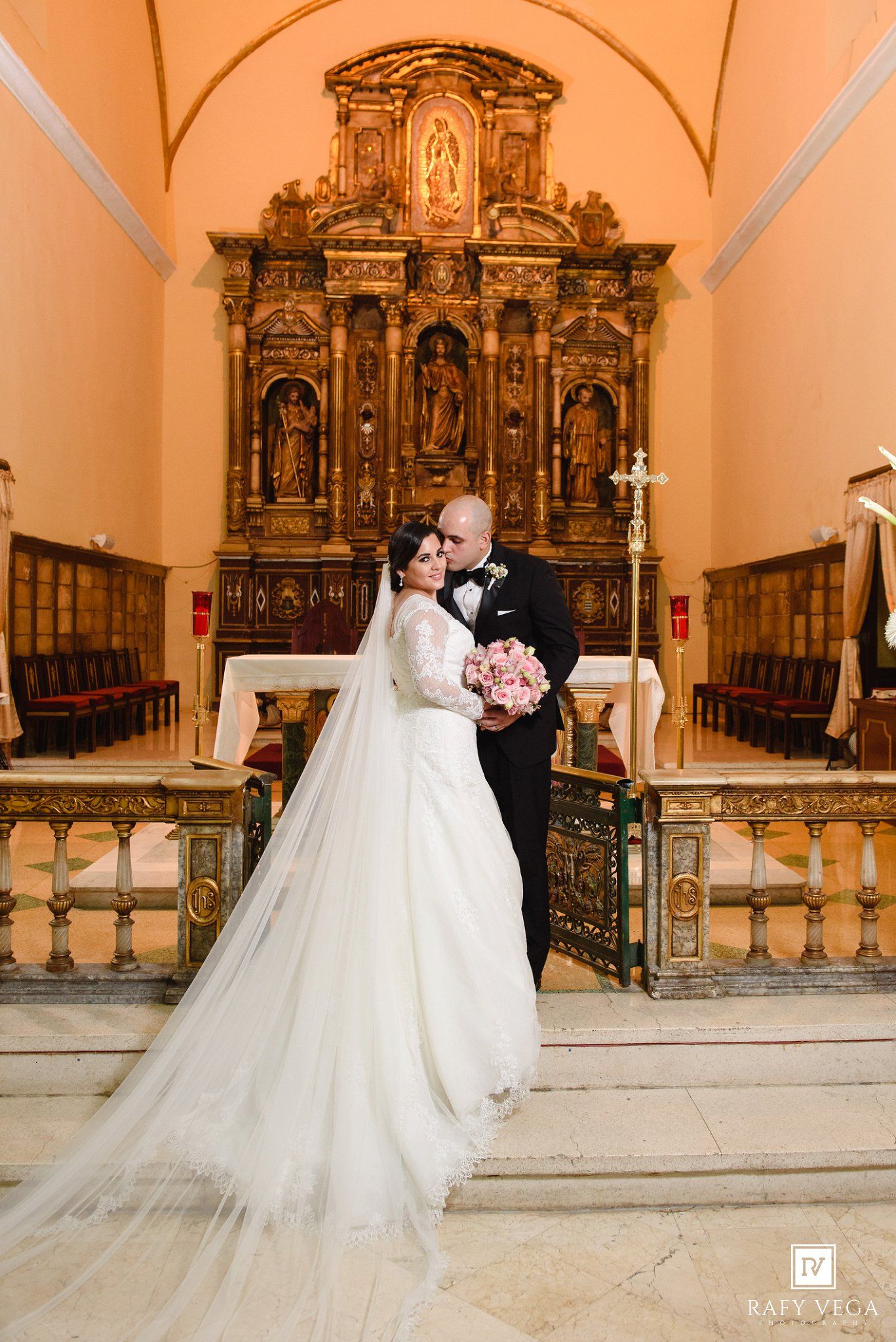 Bride and groom share a romantic moment at ornate church altar, bride in elegant gown with long veil holding pink bouquet.