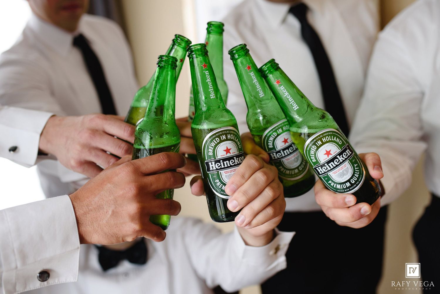 Men in white dress shirts and bow ties toasting with Heineken beer bottles at a wedding celebration.