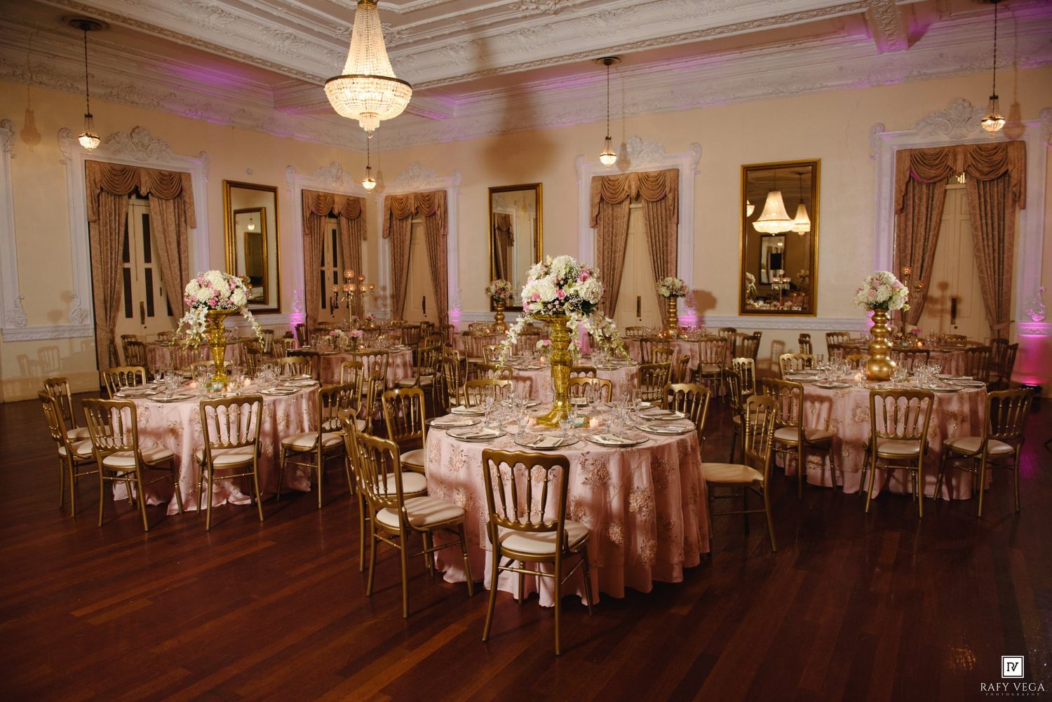 Luxurious wedding reception hall with gold chiavari chairs, pink tablecloths, and floral centerpieces.