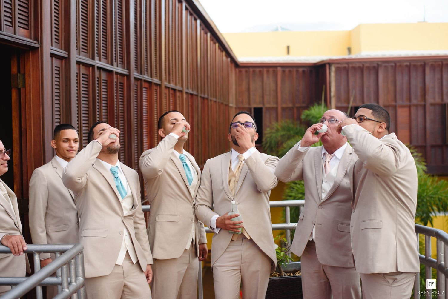 Groomsmen in tan suits toasting drinks outdoors at a wedding celebration.