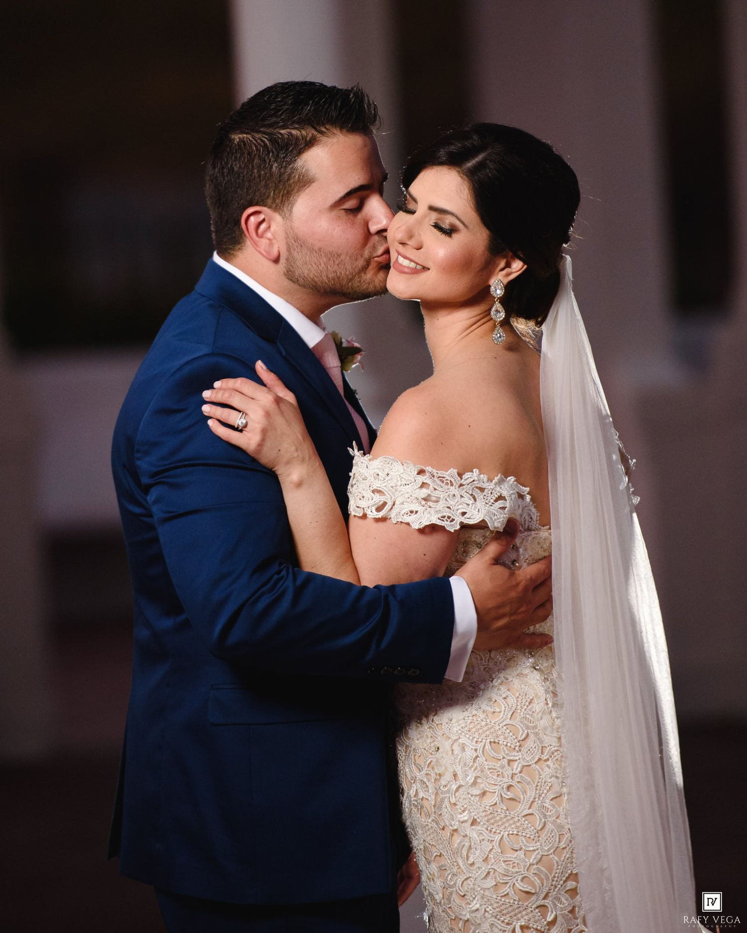 A groom in a navy suit kisses his bride in a lace off-shoulder gown and veil on their wedding day.