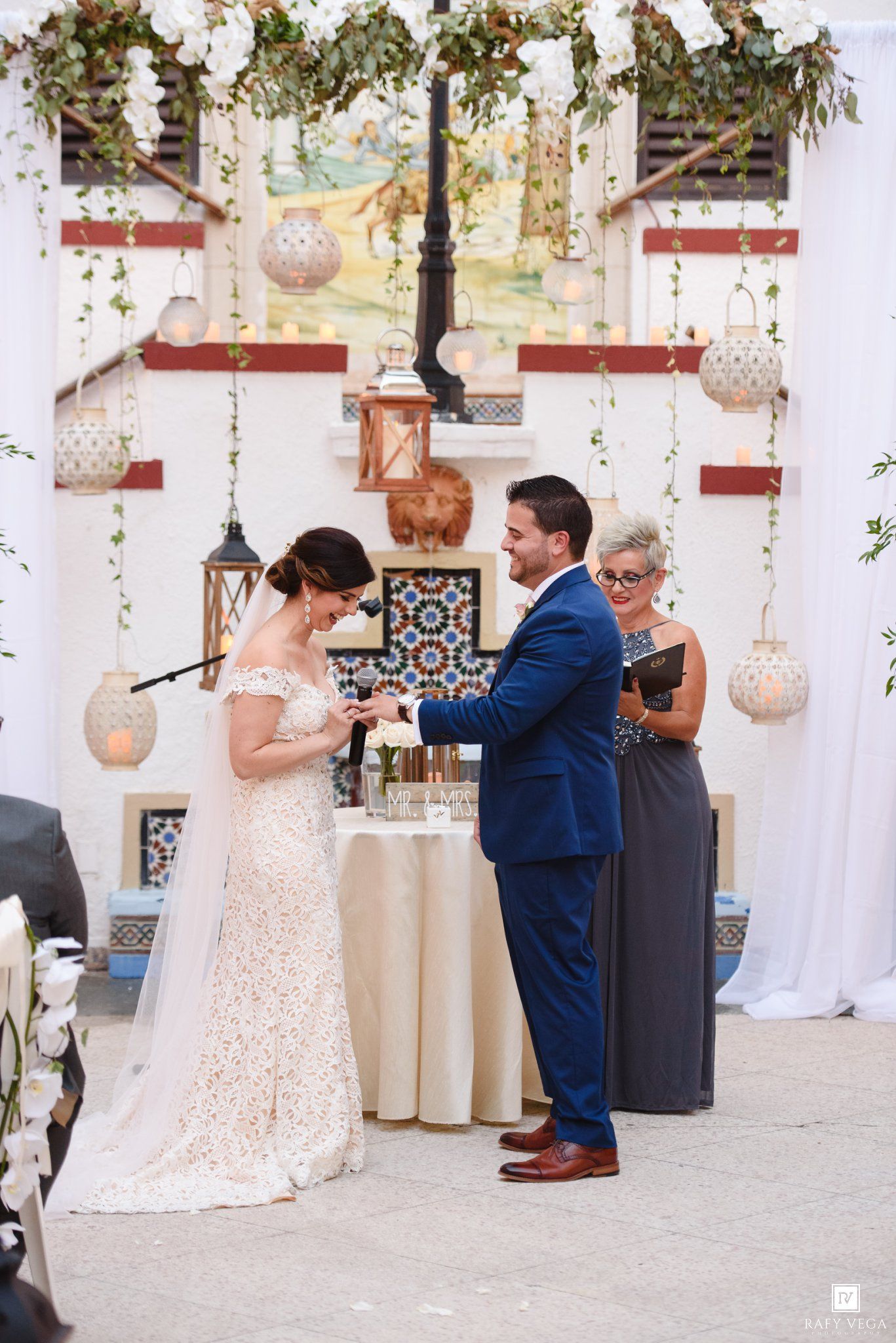 Bride and groom exchange rings during outdoor wedding ceremony with white floral decor and hanging lanterns.