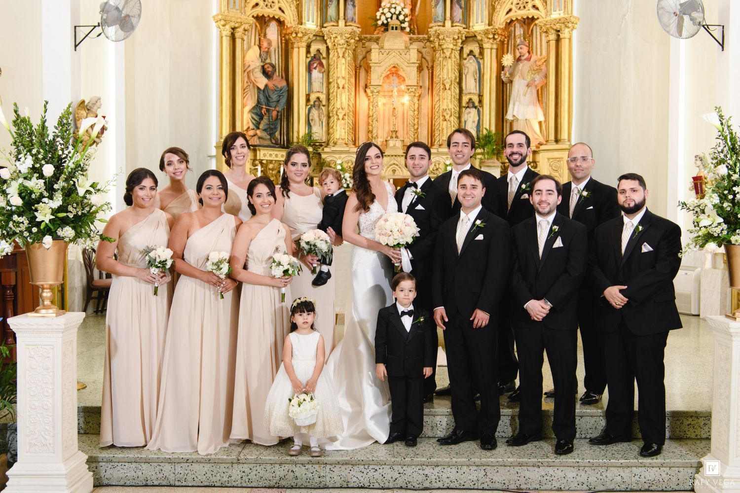 Wedding party poses in elegant church with gold altar, bridesmaids in champagne gowns, groomsmen in black tuxedos.