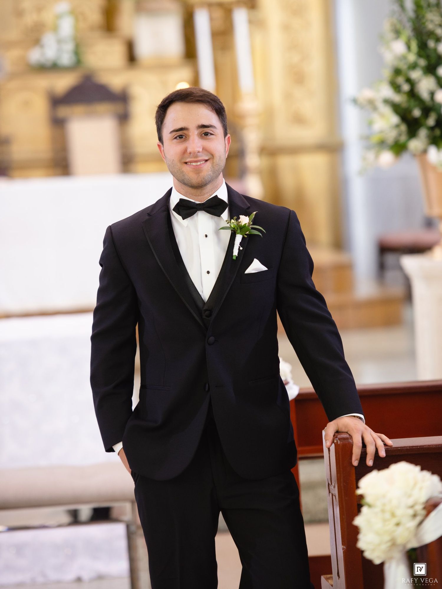 Groom in black tuxedo with bow tie and boutonniere smiling in a church before wedding ceremony.