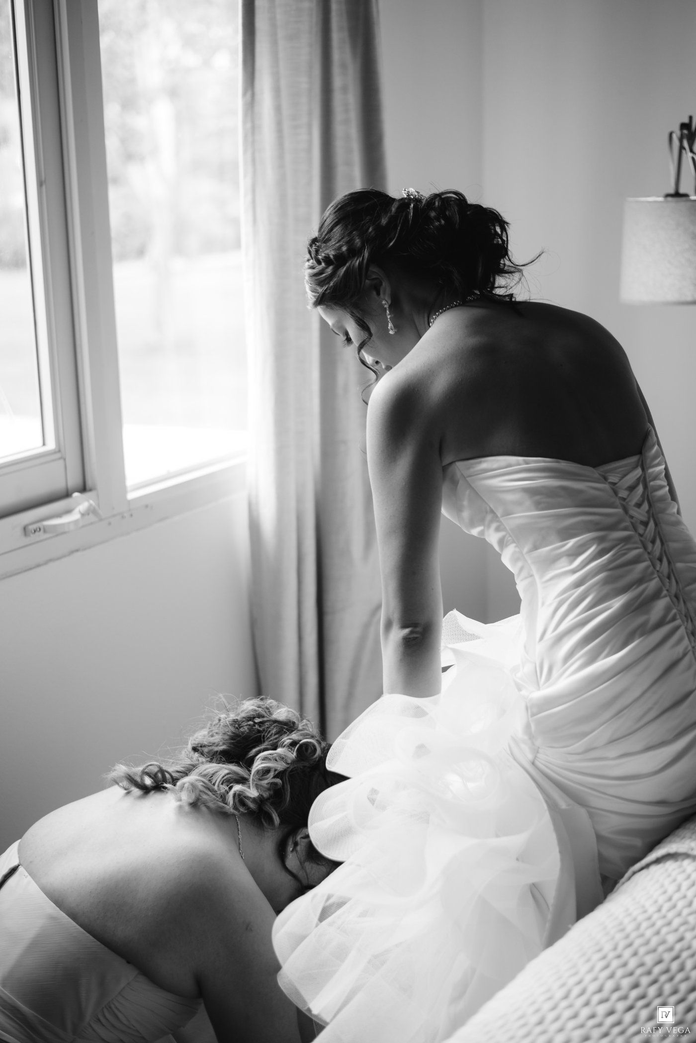 Black and white photo of a bride in a strapless gown being helped by a bridesmaid near a bright window.