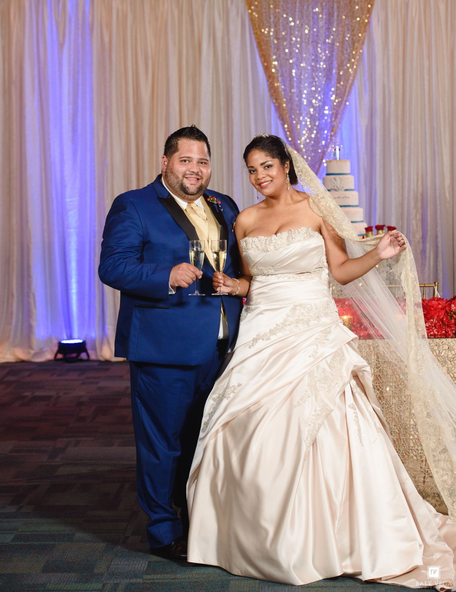 Bride in strapless satin gown and groom in blue suit toasting champagne at elegant wedding reception.