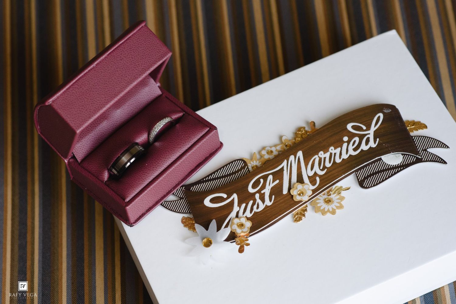 Wedding rings in a red box beside a Just Married sign with gold floral accents on a striped background.