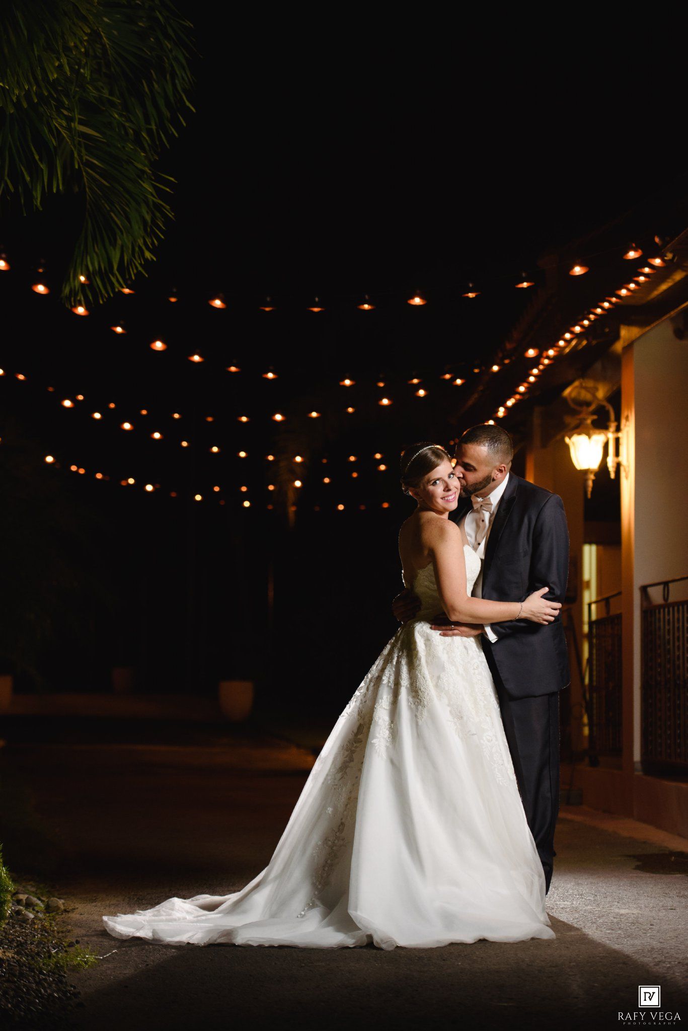 Bride and groom share a romantic embrace under glowing string lights at night during their outdoor wedding celebration.