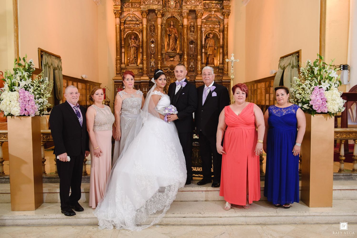 Wedding party poses in elegant church with ornate golden altar, bride in white gown flanked by family in formal attire.