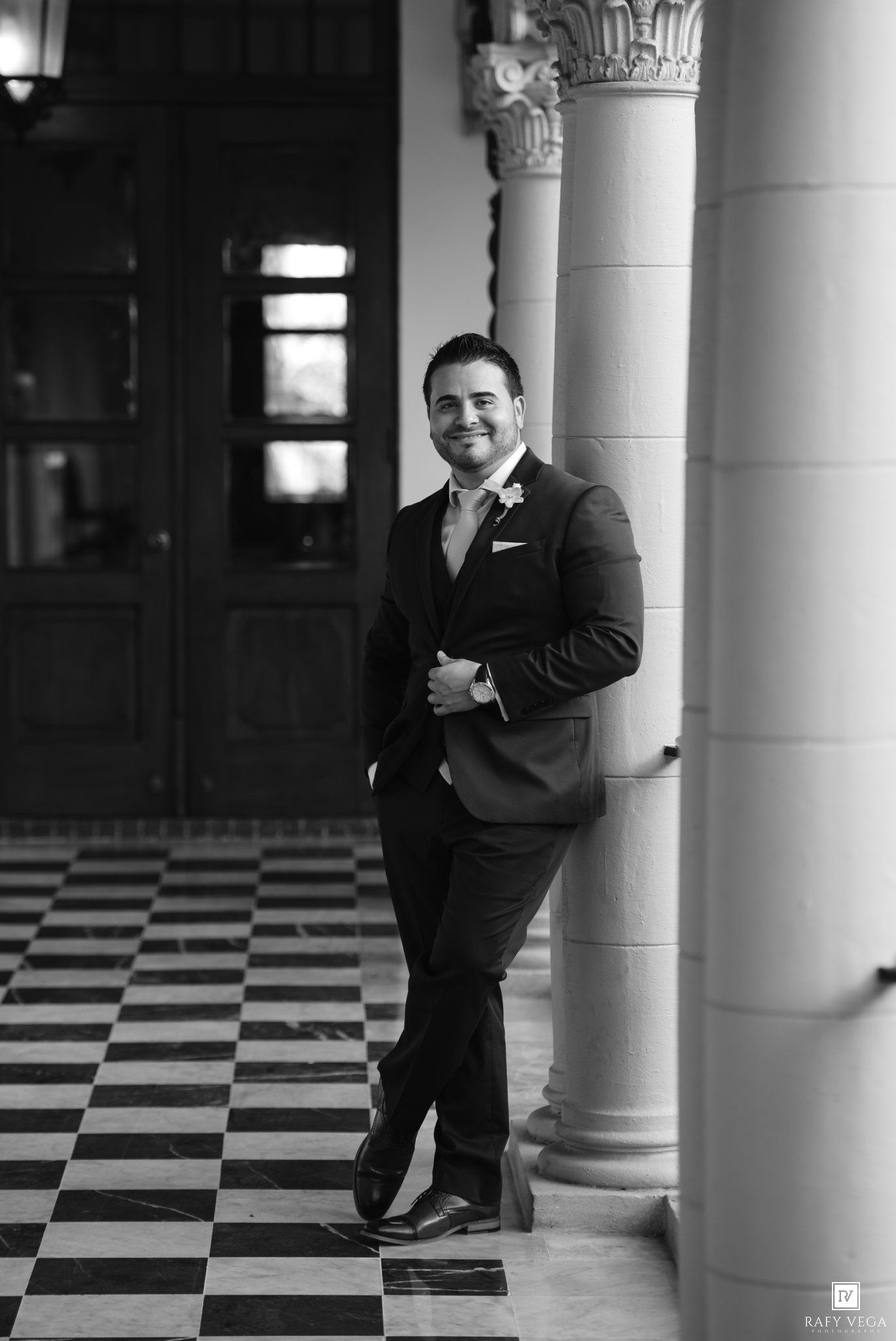 Groom in dark suit leaning against column on checkered floor in black and white wedding photo.