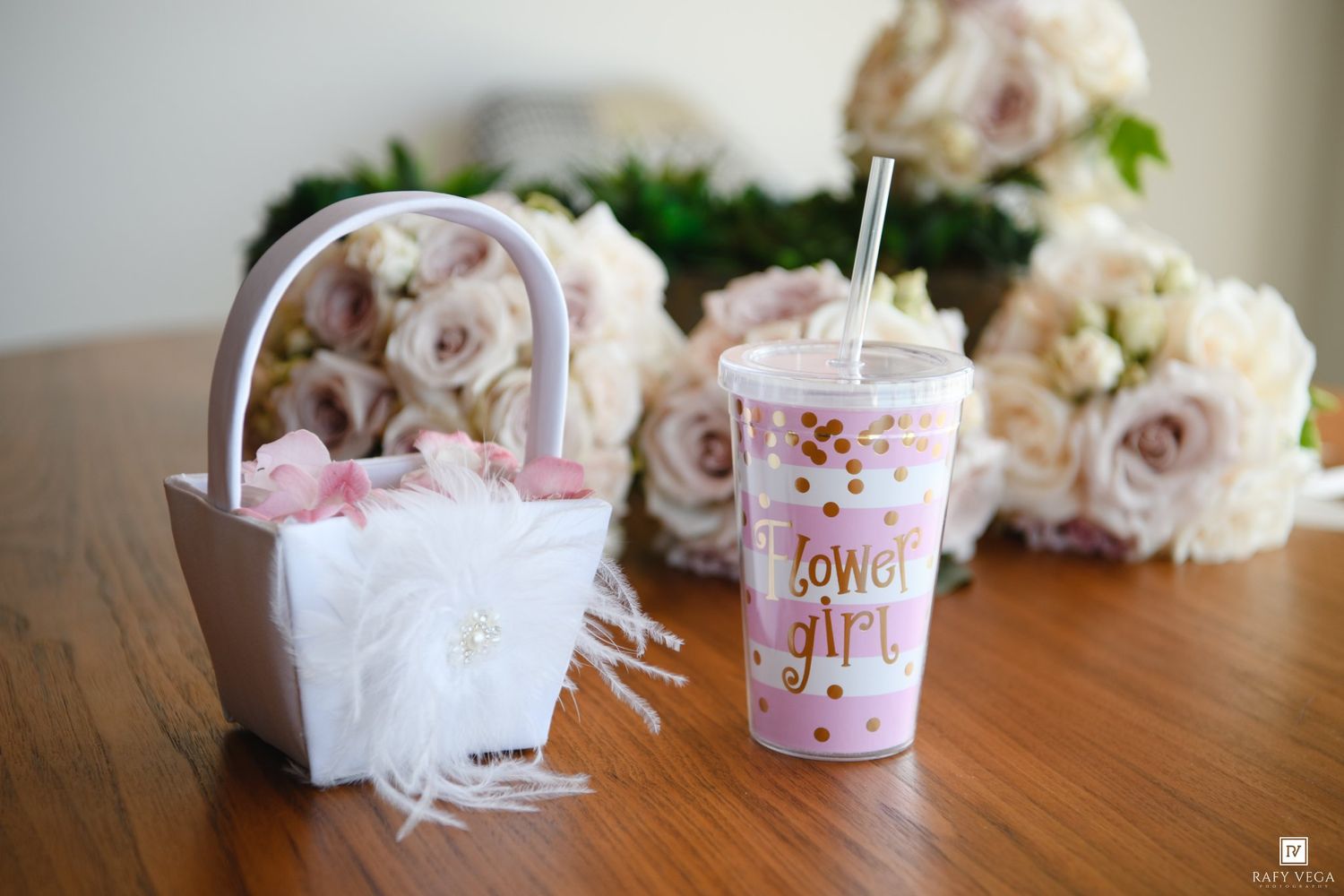 A flower girl basket and pink polka dot cup with straw on a wooden table with blush roses in the background.