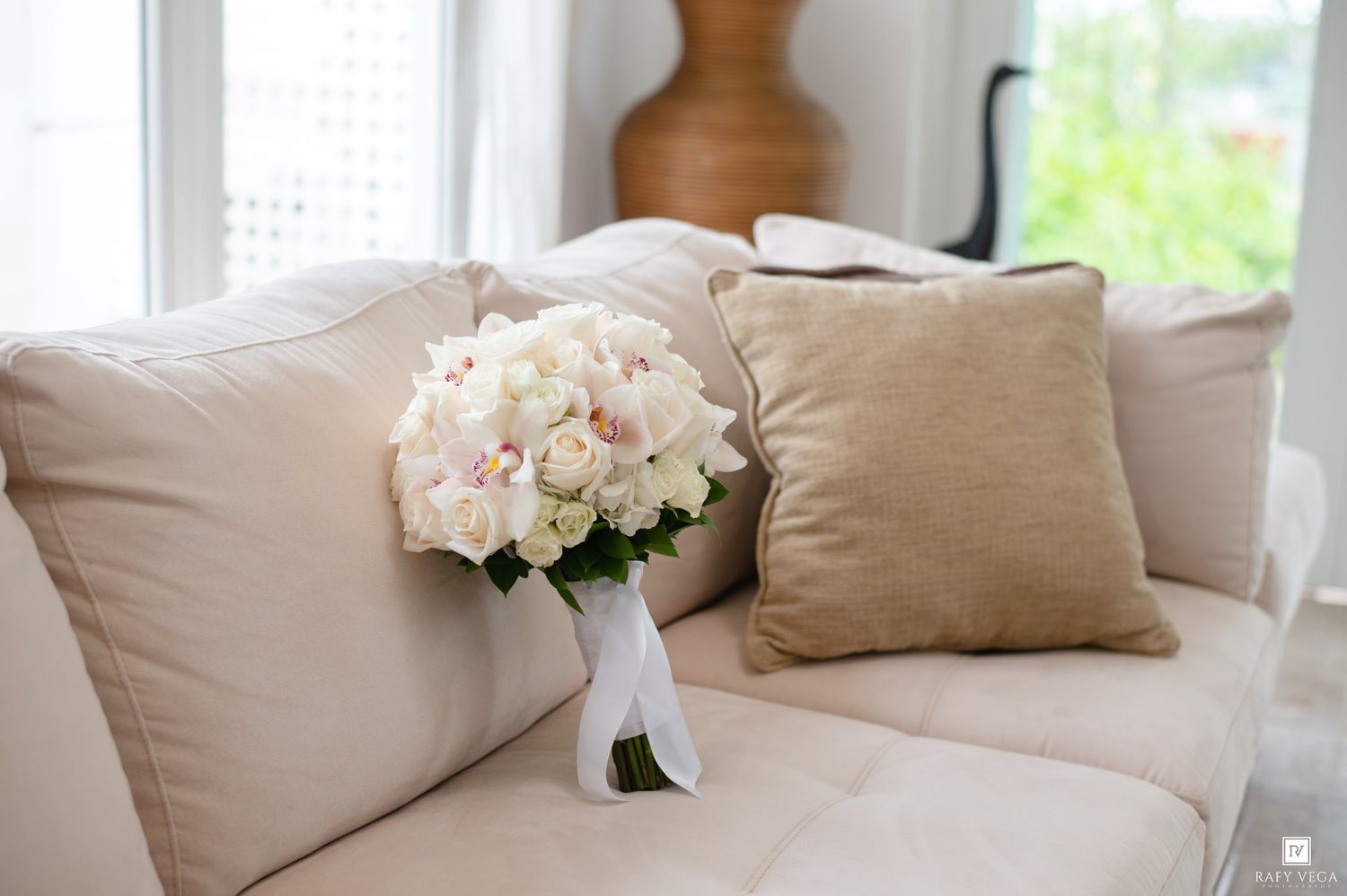 A bridal bouquet of white and blush roses rests on a cream sofa with beige pillows near a sunlit window.