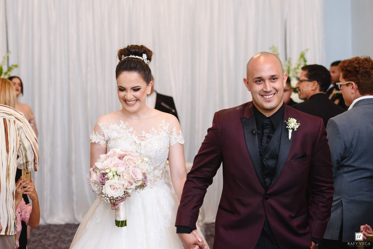 Bride and groom walk hand-in-hand after wedding ceremony, bride in lace gown holding pink bouquet, groom in burgundy suit.