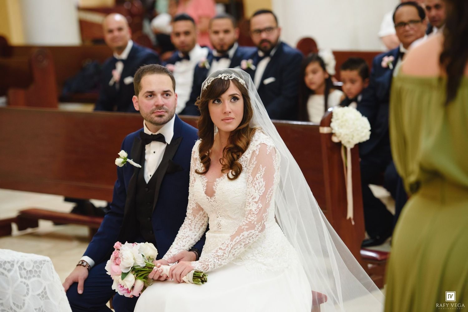 Bride and groom seated at church altar during wedding ceremony, bride in lace gown holding pink bouquet.