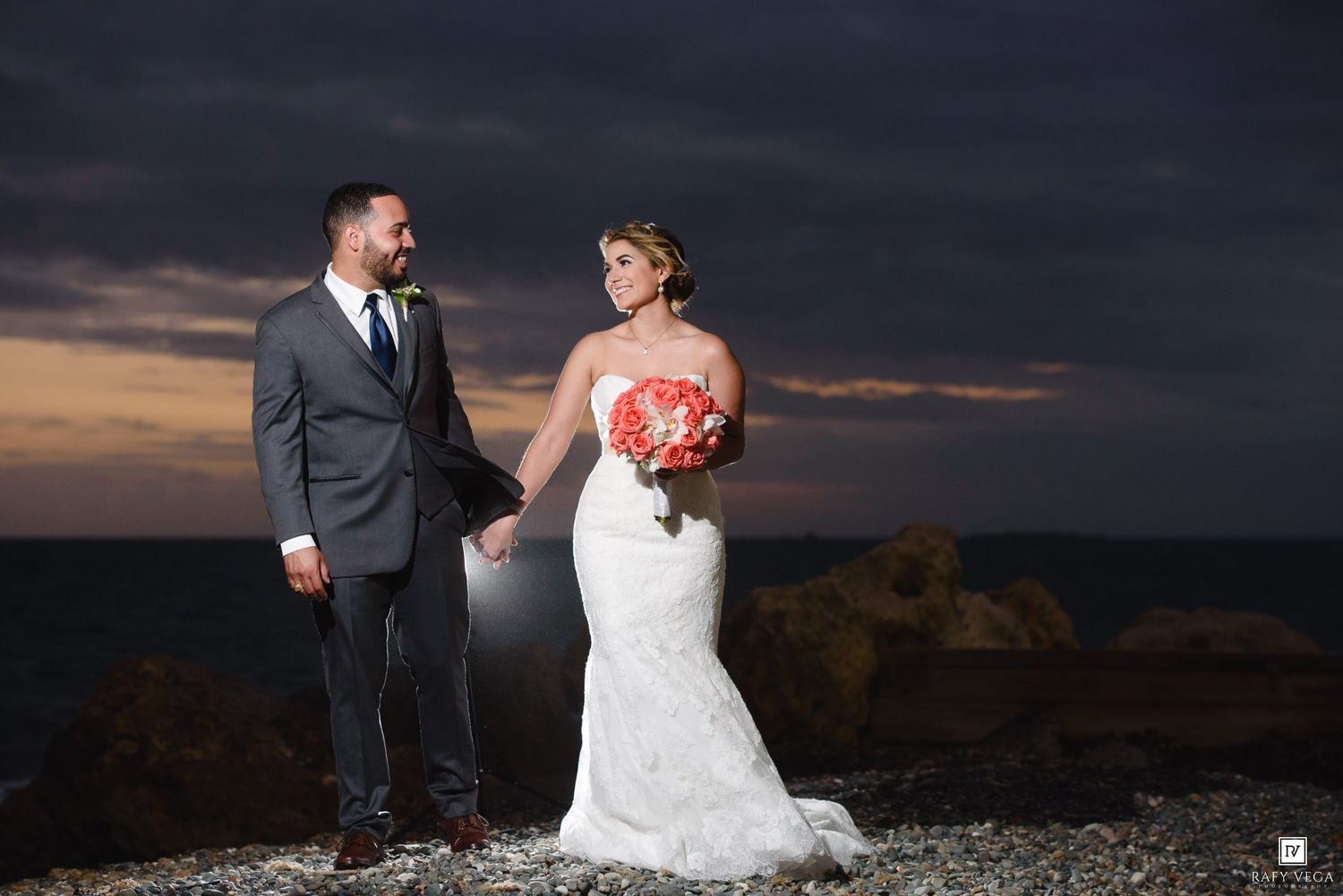 Bride and groom holding hands at rocky ocean shore at sunset, bride in lace gown holding red bouquet.