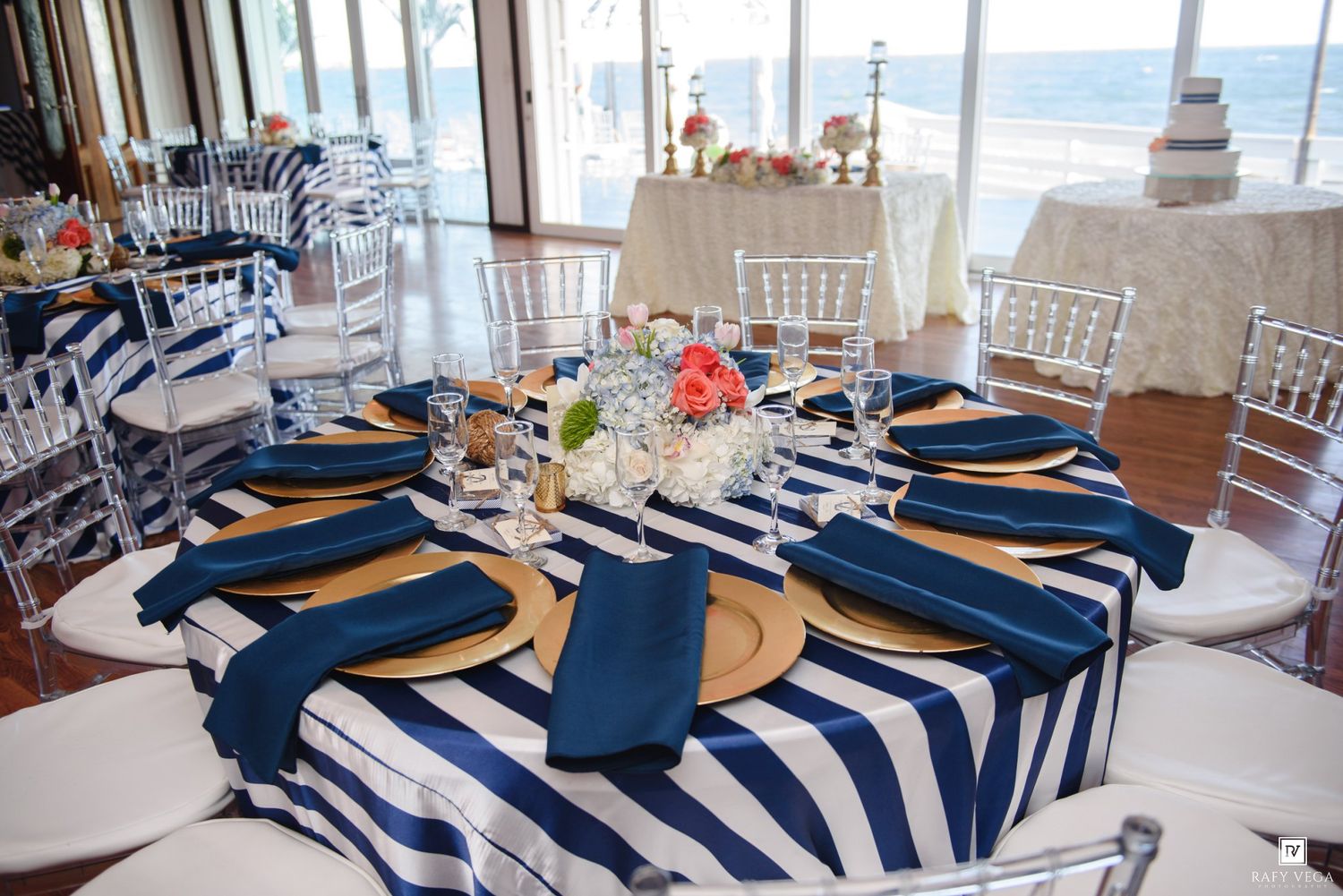 Navy blue and white striped tablecloths with gold chargers and blue napkins set for a beachfront wedding reception.