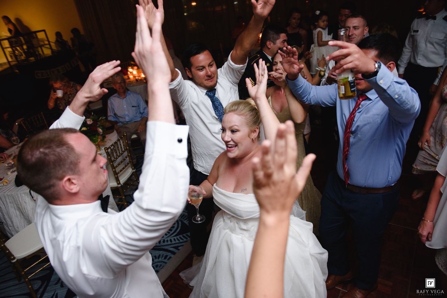 Bride and groom celebrating with raised hands surrounded by cheering guests at wedding reception.