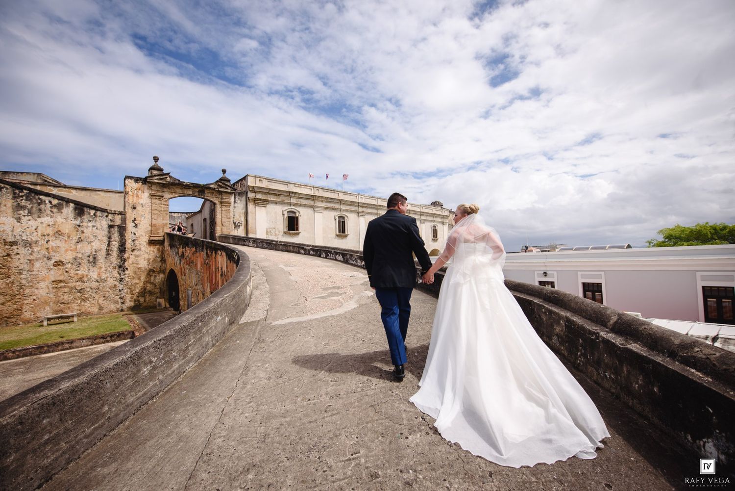 Bride and groom walk hand-in-hand along a historic stone fortress path under a dramatic cloudy sky.