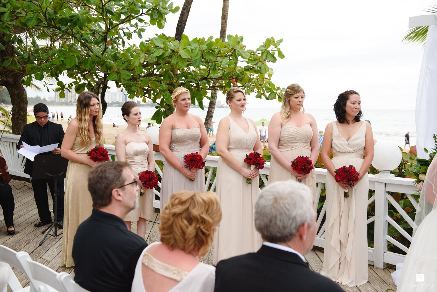 Bridesmaids in champagne gowns holding red bouquets stand at an outdoor beach wedding ceremony.