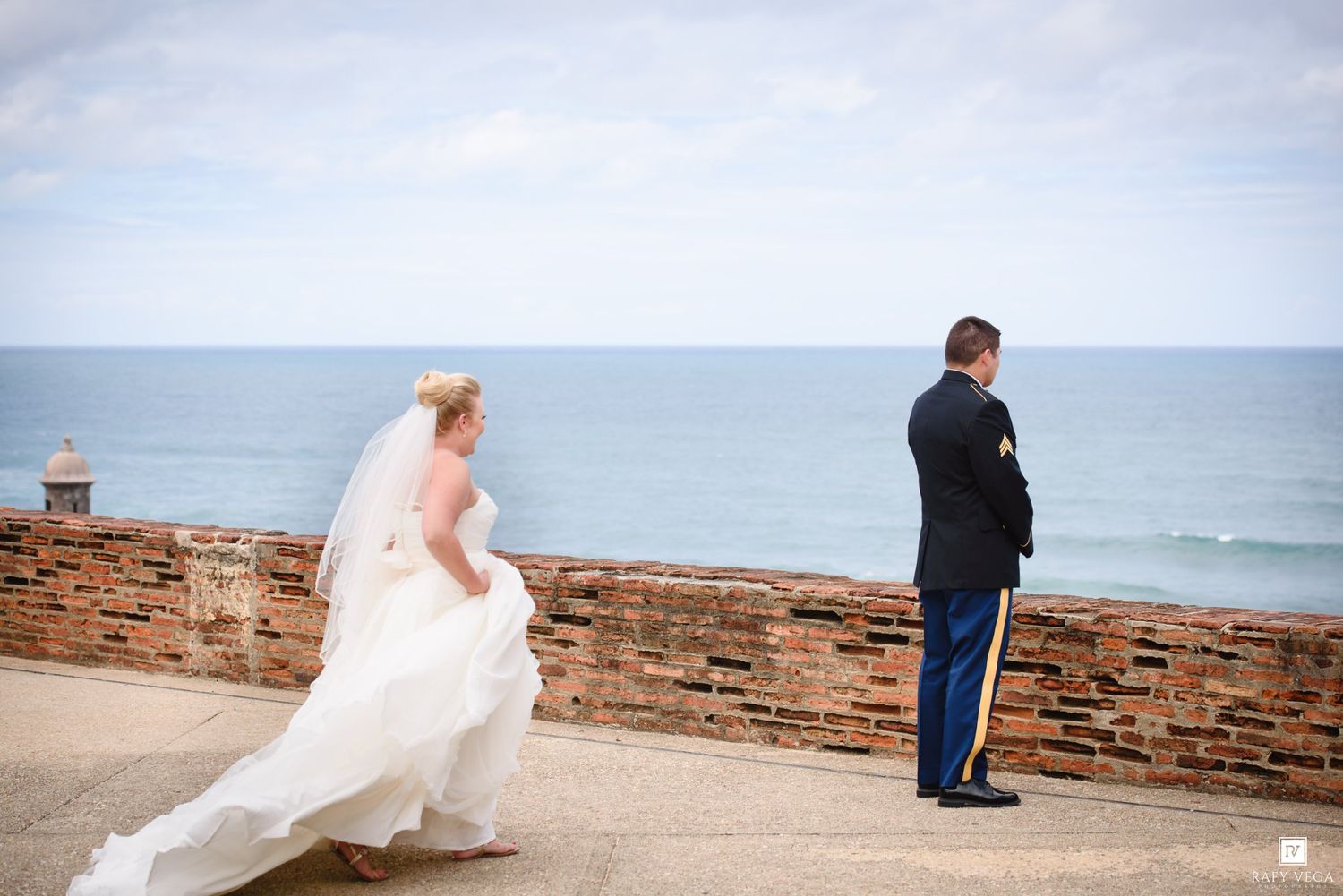 Bride in white gown and veil approaches groom in military uniform for first look by ocean wall.