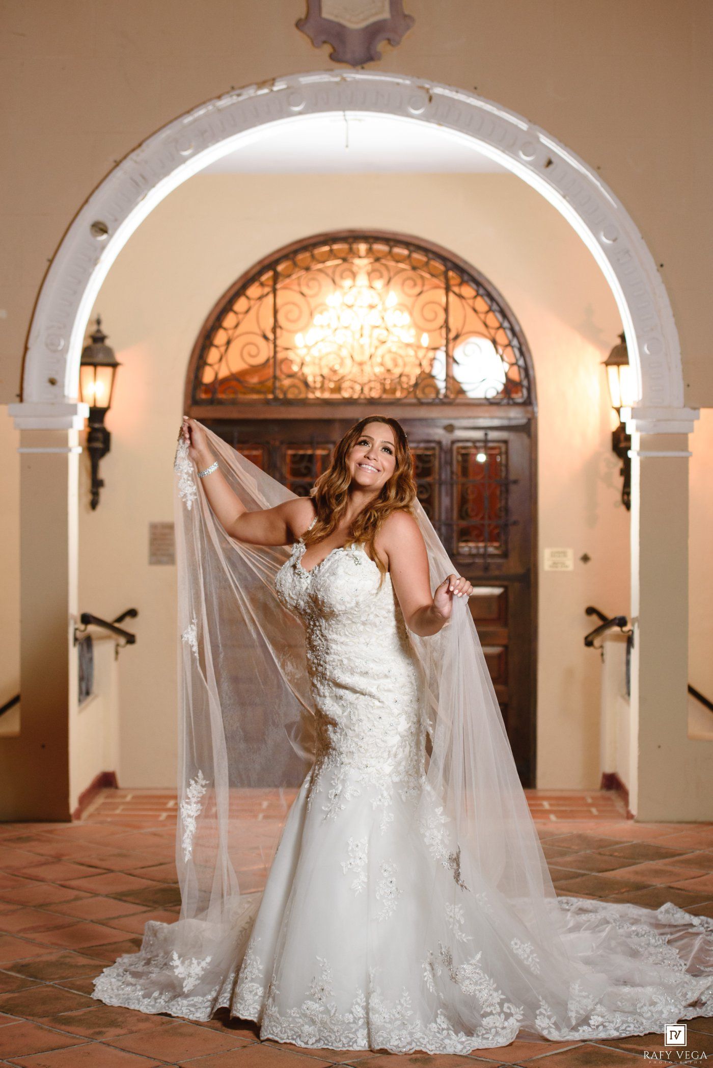 Bride in lace mermaid wedding gown holding flowing veil under elegant arch at luxury venue.
