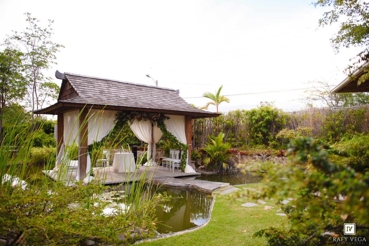 Elegant outdoor gazebo with white curtains and greenery beside a serene pond in a lush tropical garden setting.