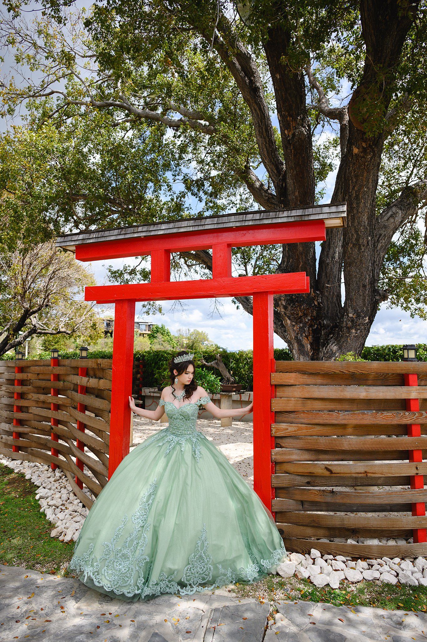 Young woman in mint green quinceañera ball gown poses under a red torii gate surrounded by lush trees.
