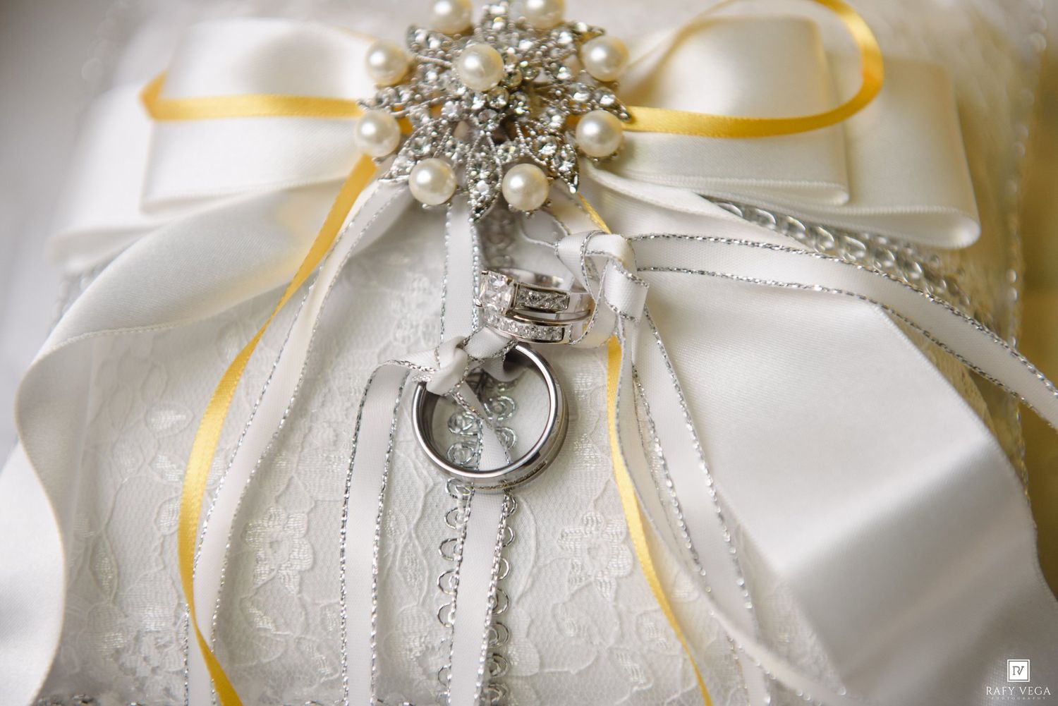 Wedding rings on white lace pillow with pearl brooch and yellow ribbon decoration.