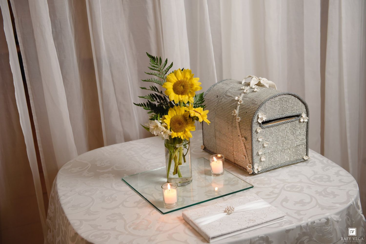 Elegant wedding gift table with sunflowers in a vase, silver card box, candles, and white lace tablecloth.